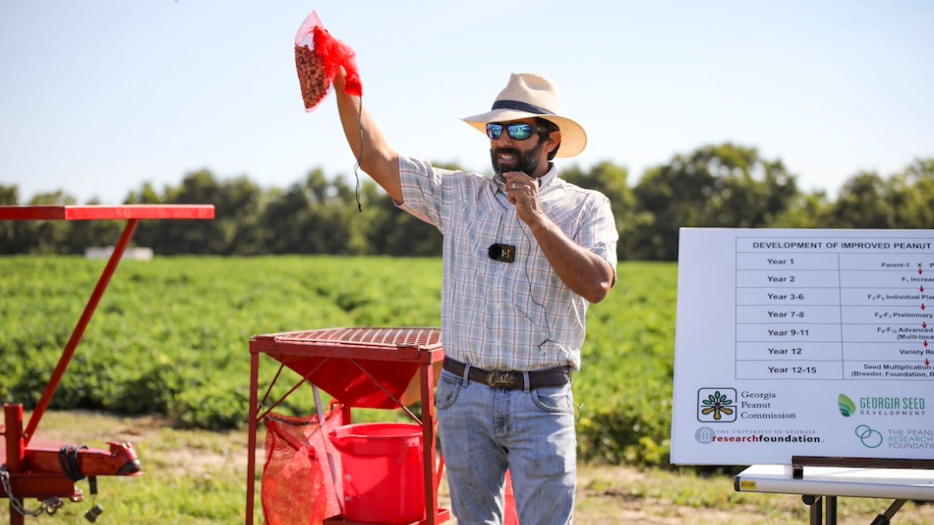An individual holding a red bag of peanuts and speaking into a handheld microphone in an agricultural setting.