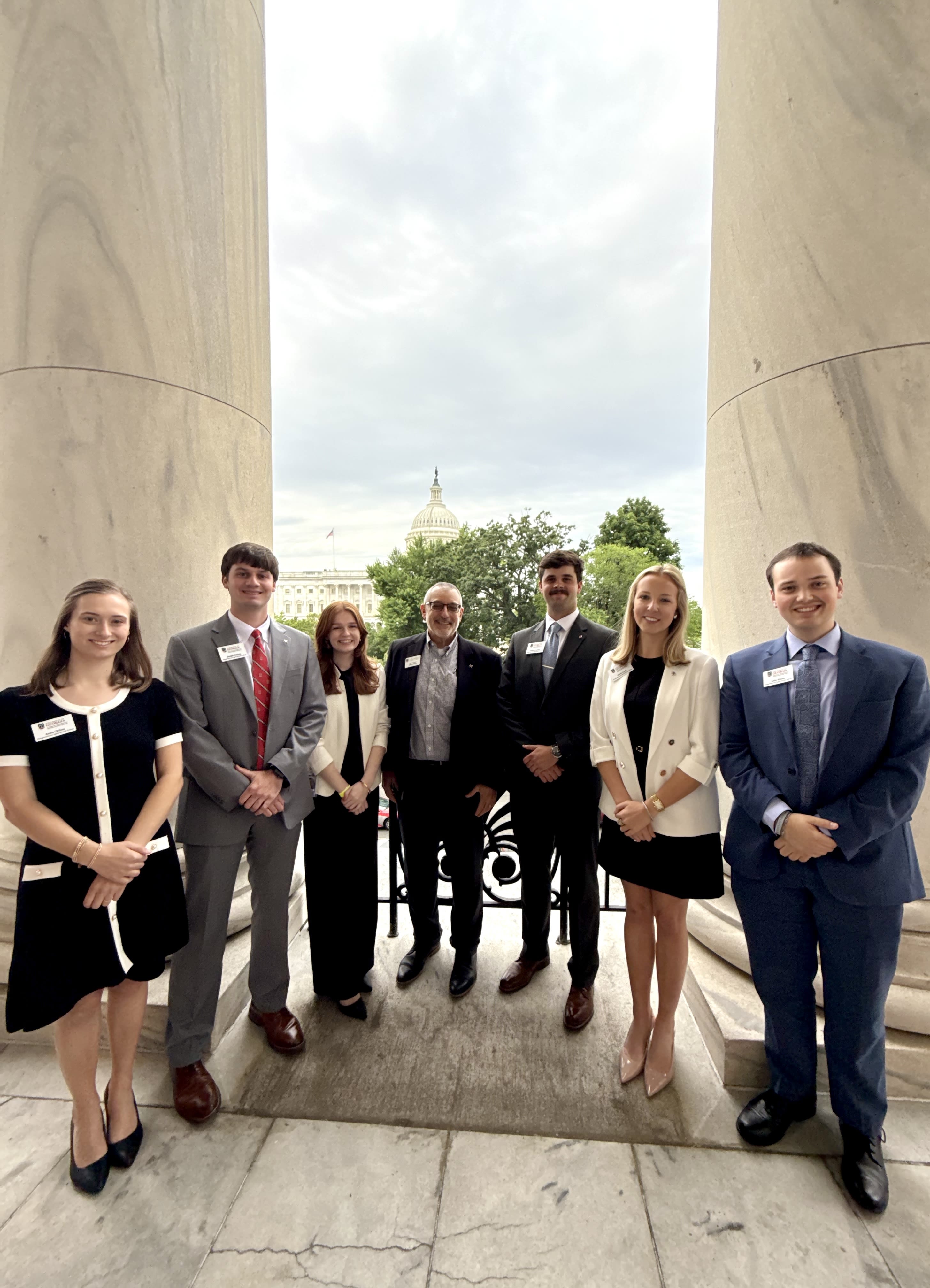 Seven individuals in formal dress standing between two large white columns with the United States Capitol Building in the background.