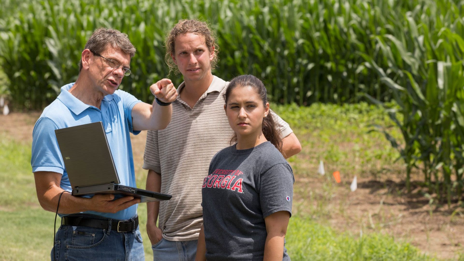 Three individuals standing in a farm field. One holds a computer.