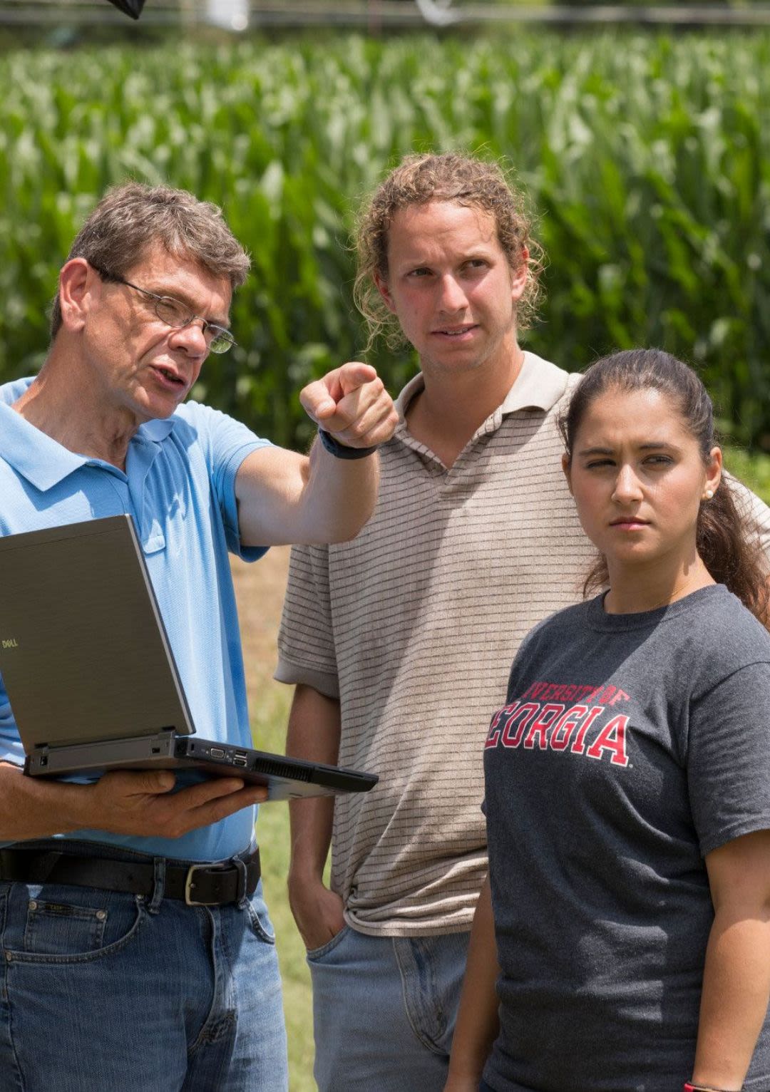 Three individuals standing in a field. One holds a computer.