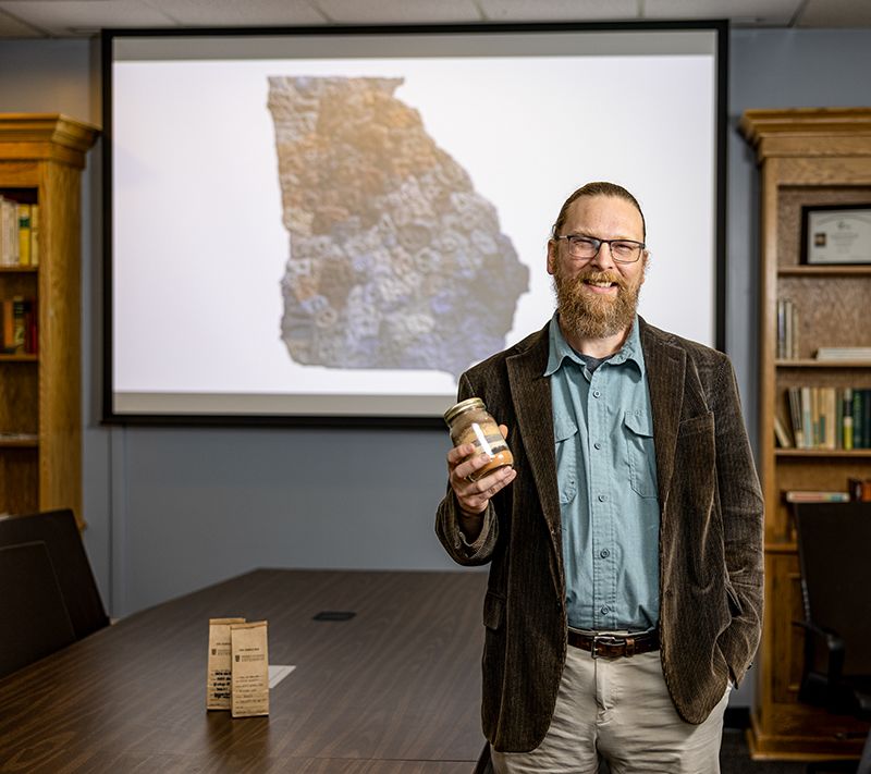 A man holds a jar of striated dirt in front of a screen displaying a dirt map of Georgia