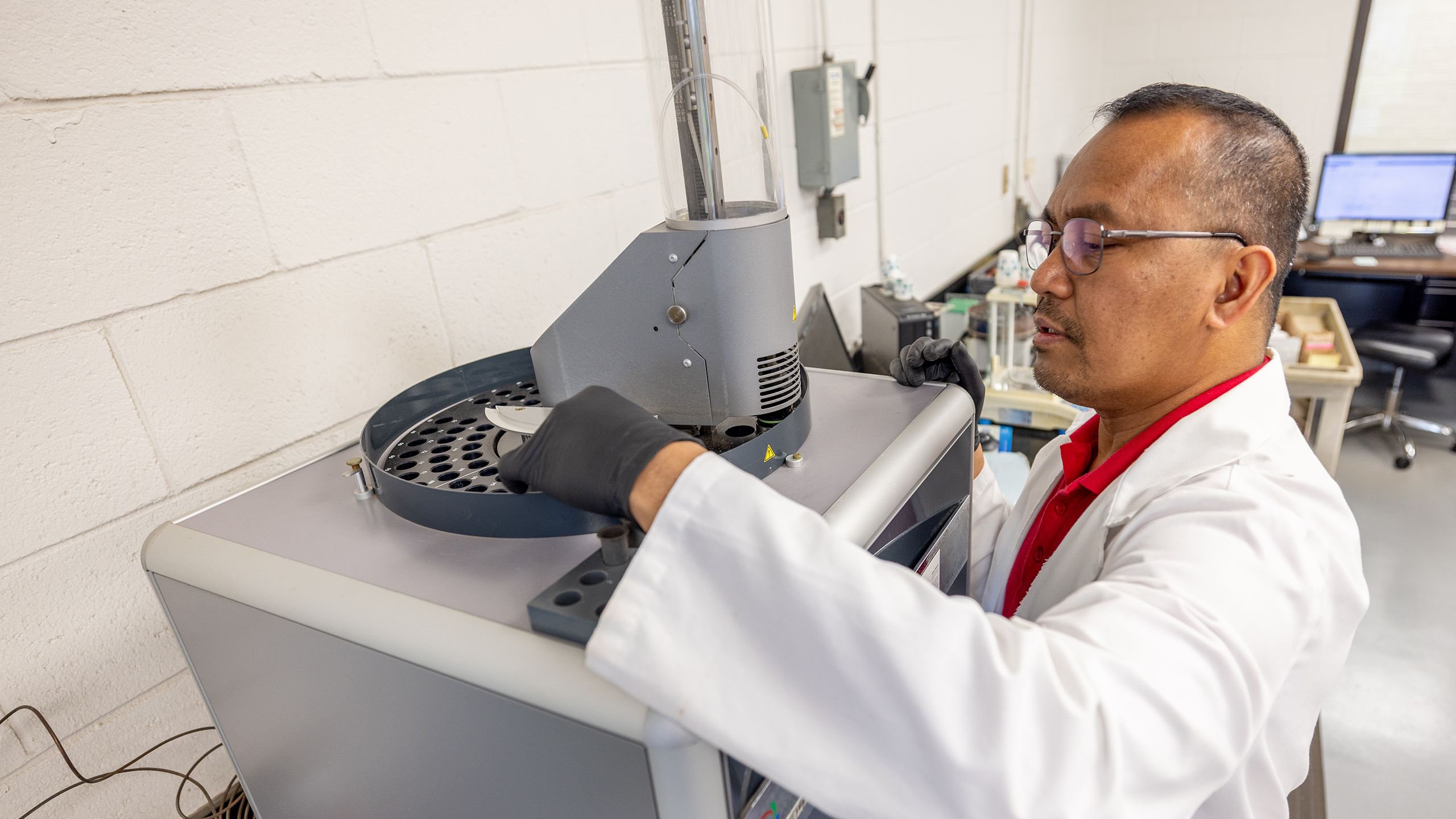 A man wearing a lab coat puts small vials into a round tray atop a machine.