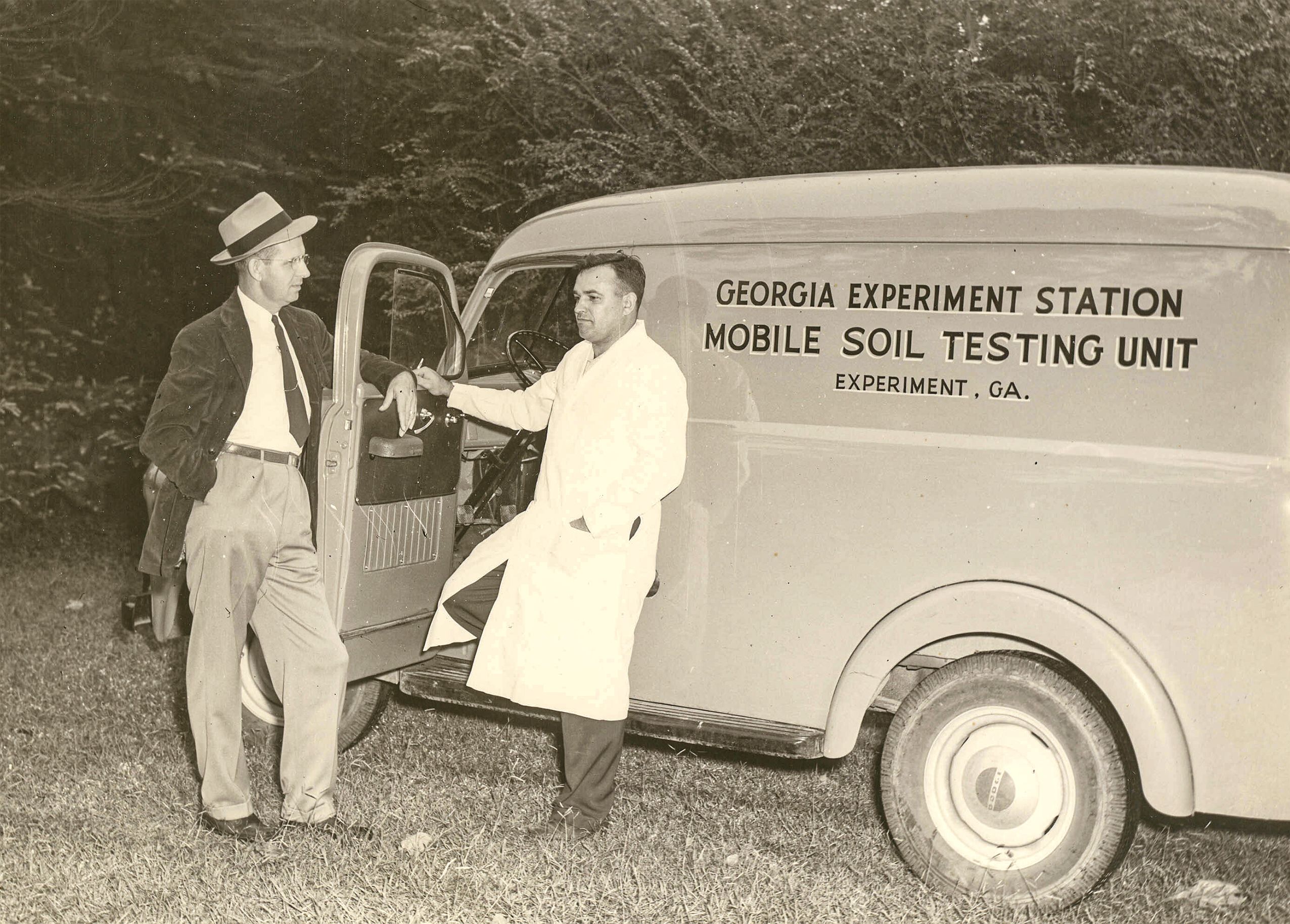 1950s era black and white photo of two men leaning on the door of a soil testing truck. One man is wearing a hat, suit and tie. The other man is wearing a lab coat. 