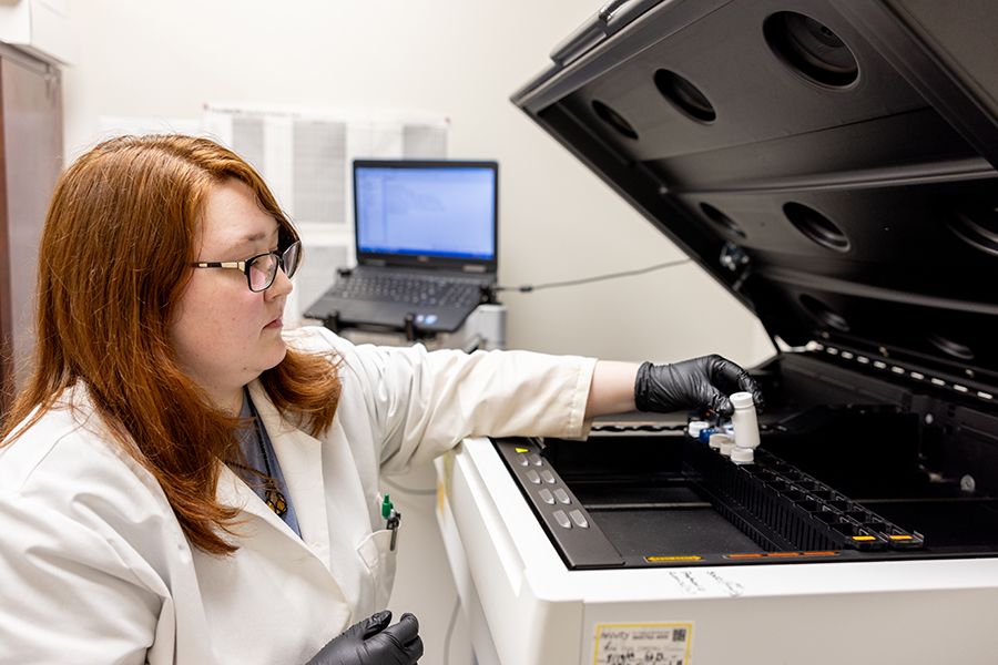 A young woman puts a small white container into a large machine for testing.