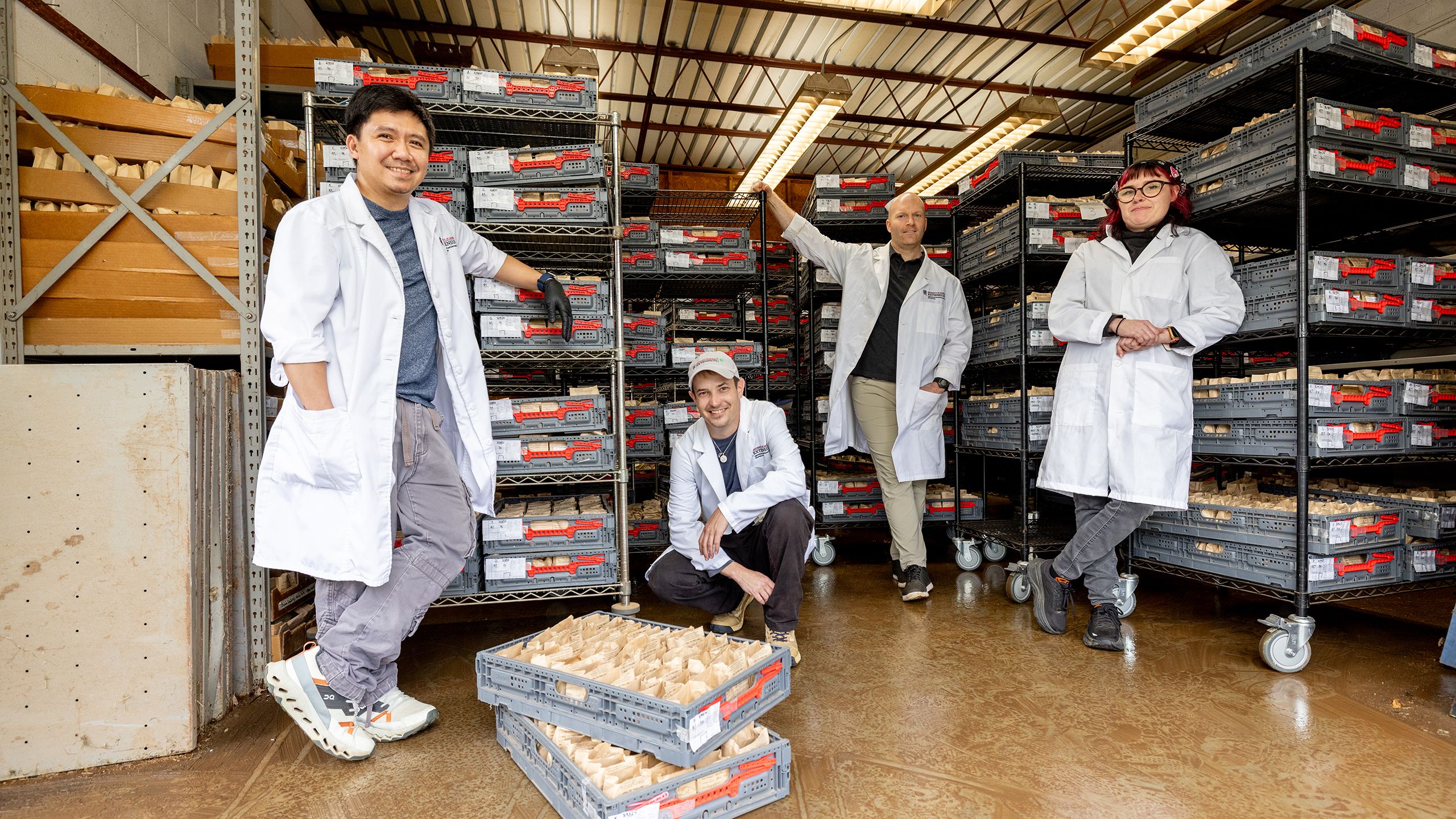 Four researchers wearing lab coats stand and kneel among shelves filled with trays of labeled paper soil sample bags in a storage room.
