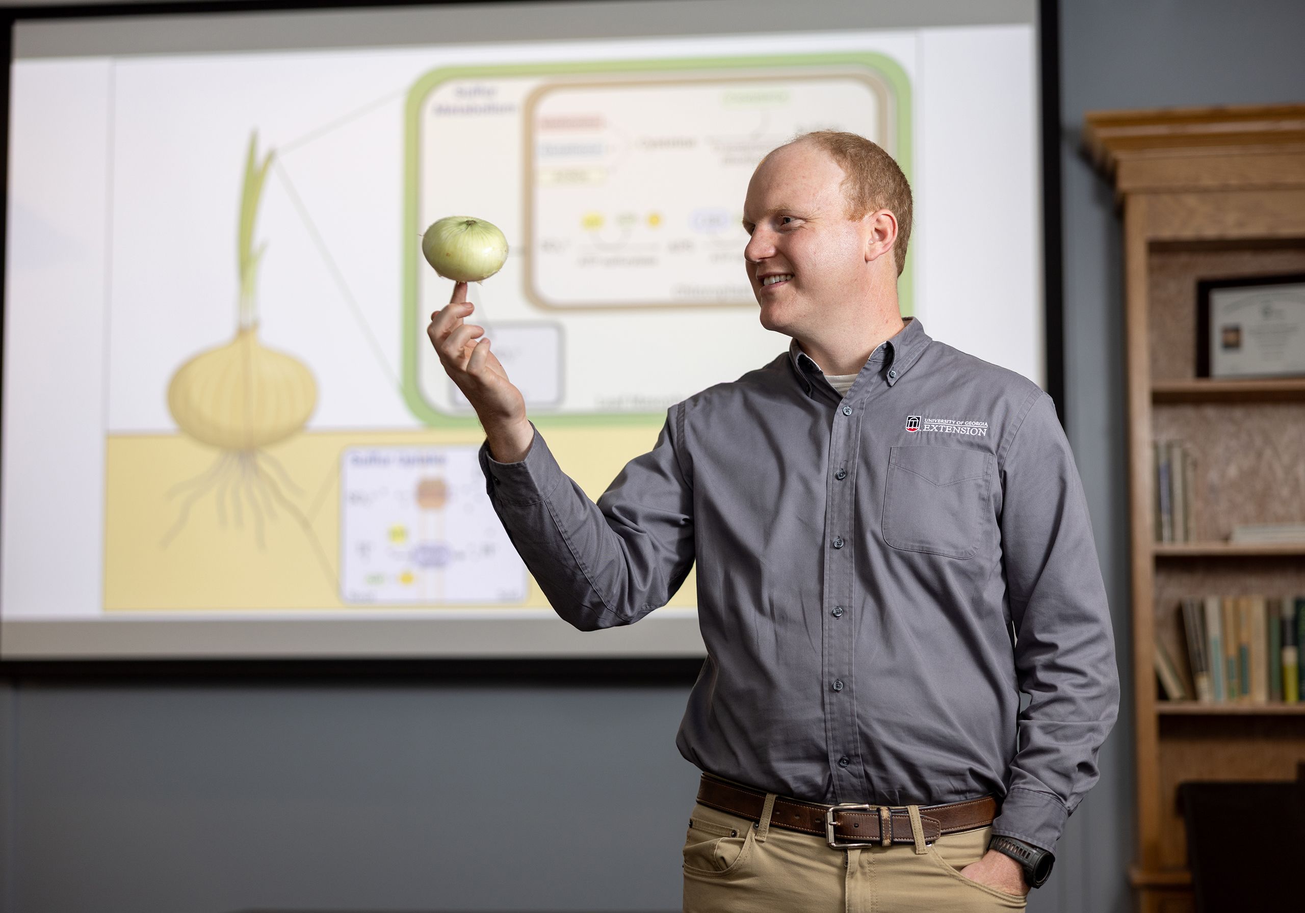 A man balances an onion on his finger tips. Behind him, an infographic shows an onion on a large screen.