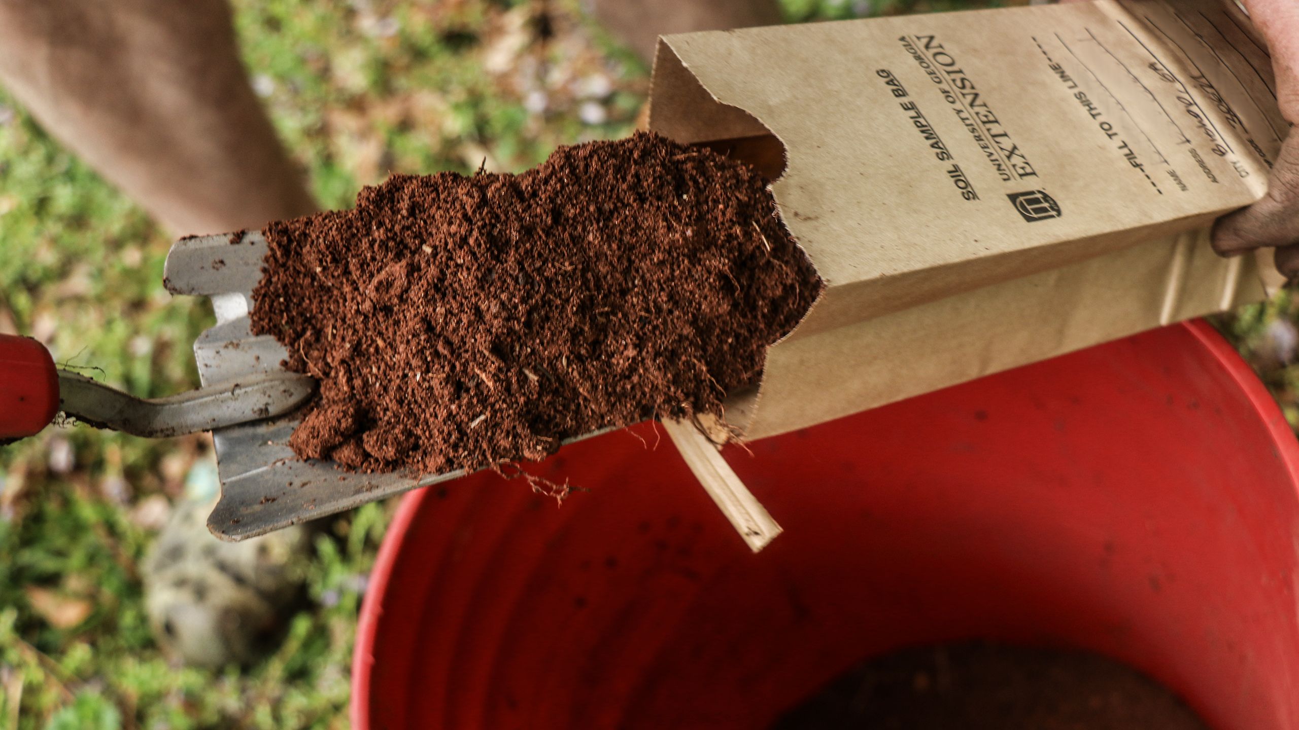 Trowel scoops red dirt from a bucket into a soil sample bag