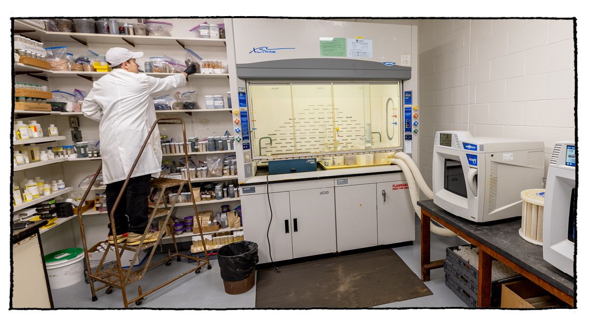 A man in a lab coat stands on a ladder to retrieve a sample from a wall of shelves. A vent hood and other lab equipment is to his right.