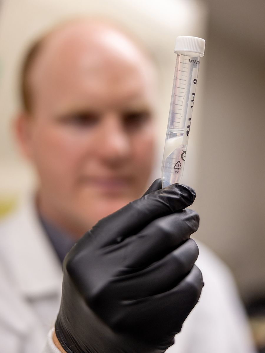 A gloved man holds a small vial containing a clear liquid and white plant material.