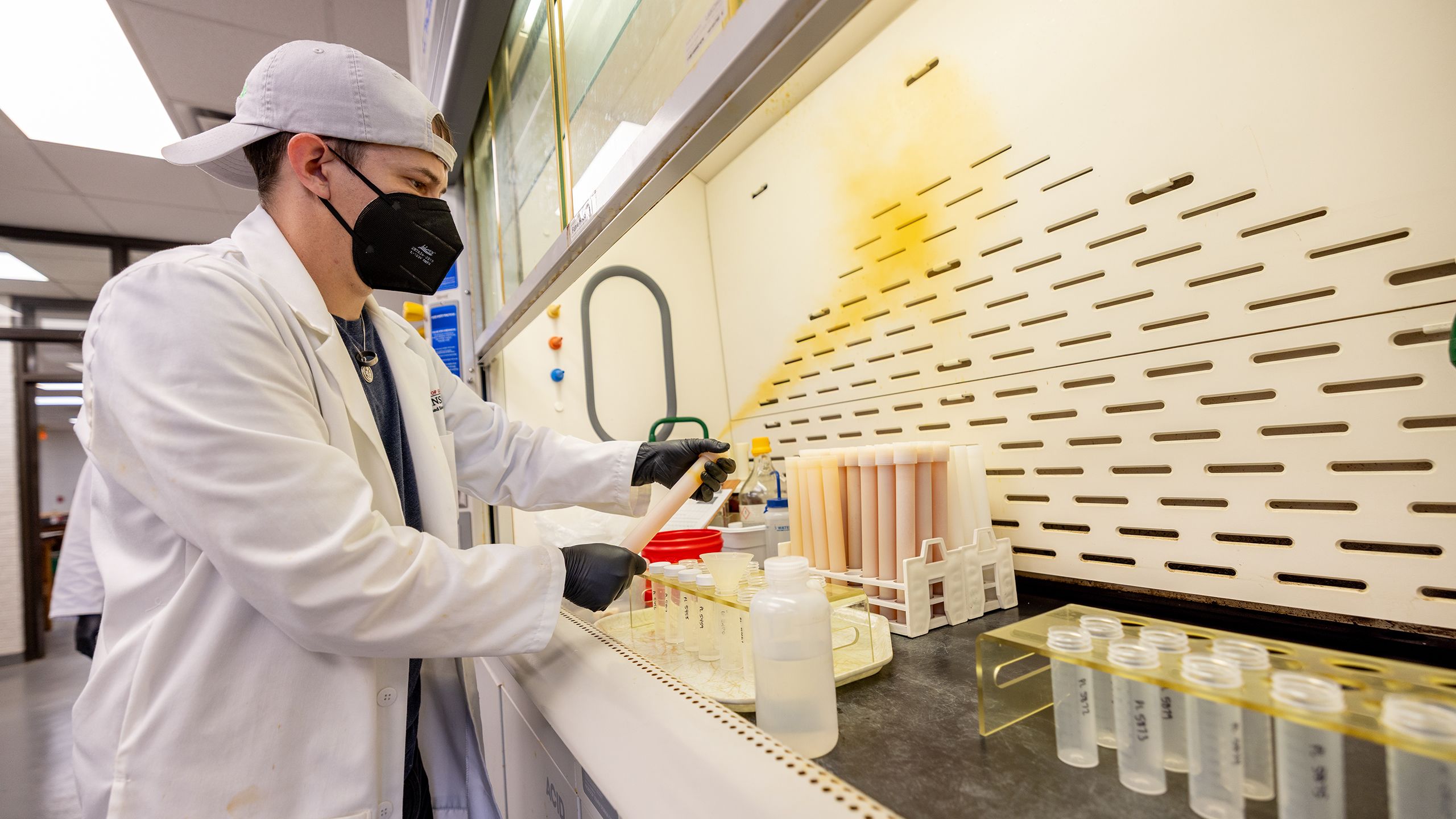 A man in a lab coat, gloves and face mask slowly opens a pressurized tube inside a vent hood. A plume of orange steam is emitted from the sample.