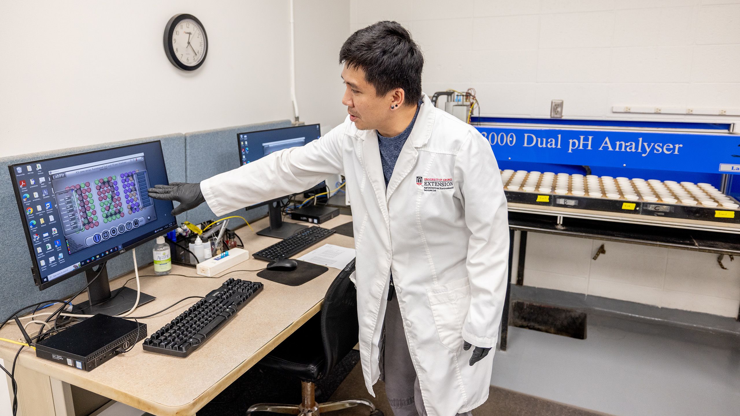 A man in a lab coat gestures to a screen in front of a large machine analyzing large trays of samples in paper cups