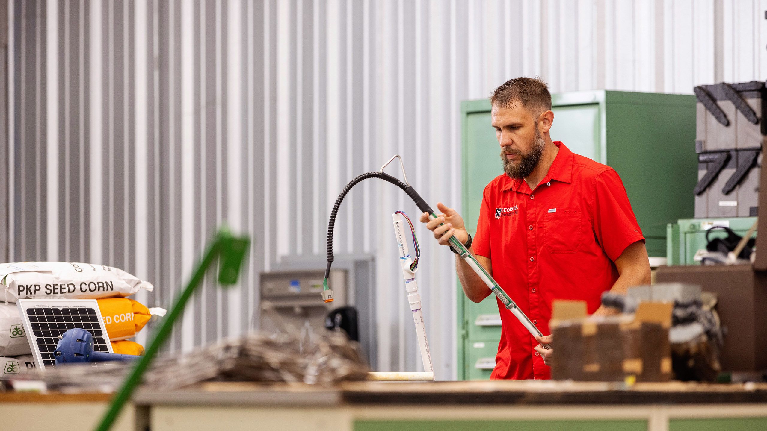Man in a red shirt examines a long tool with wires in a workshop filled with equipment and seed corn bags.