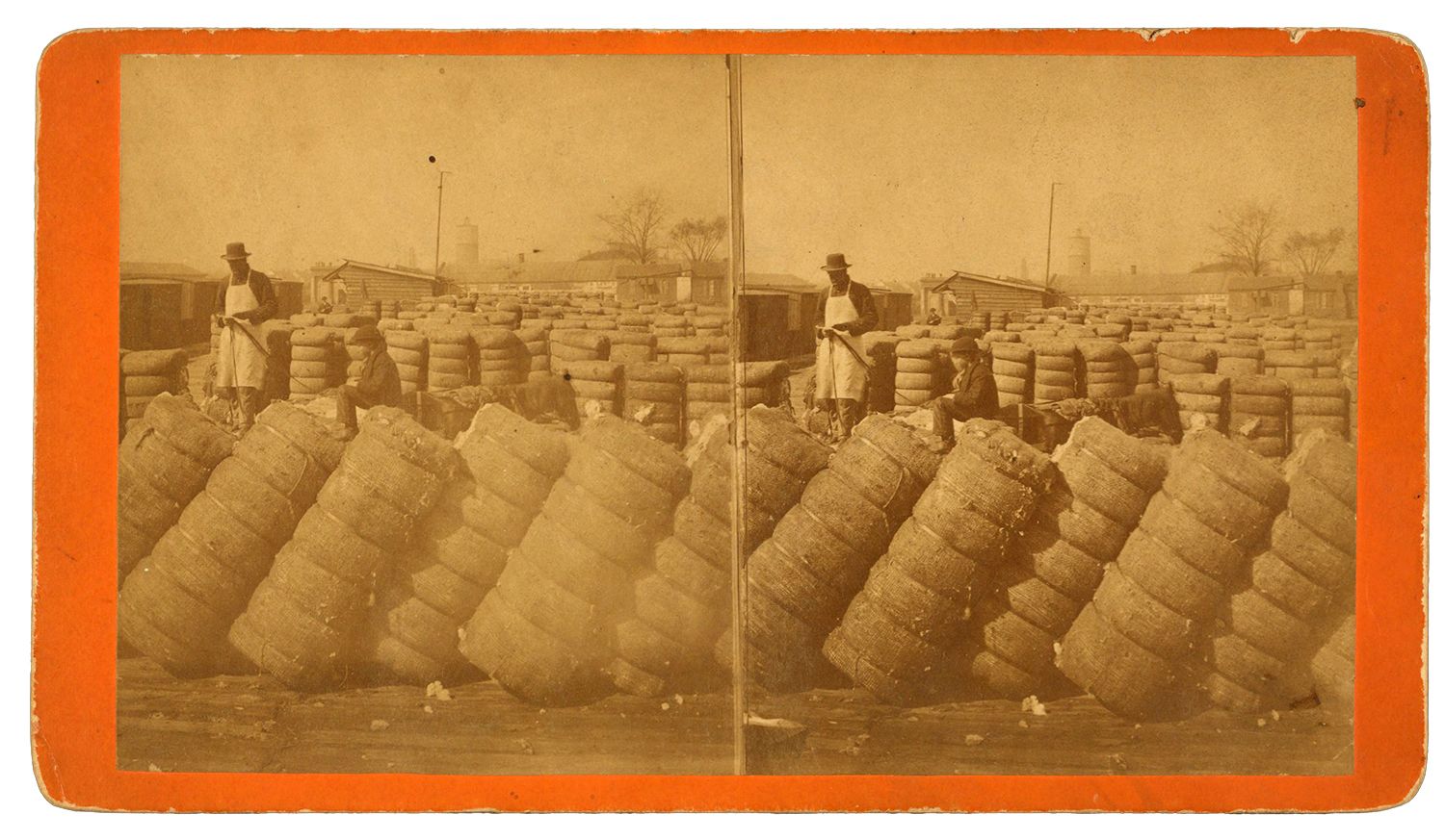 Men stand among stacked cotton bales in a large outdoor yard, with buildings and bare trees in the background.