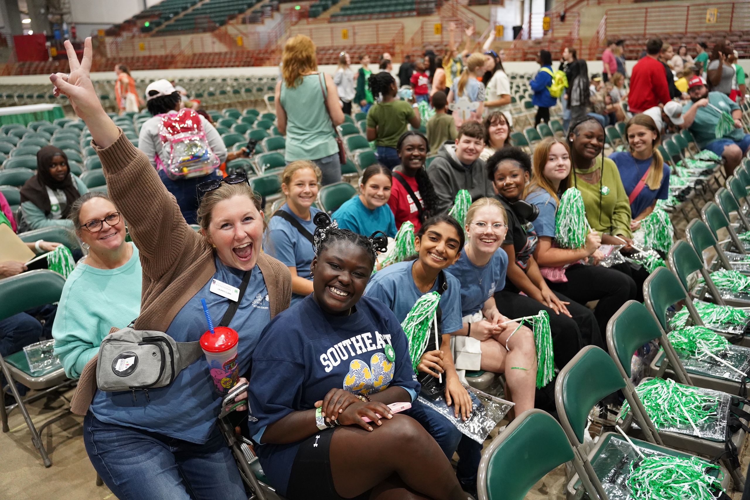 Group of people in blue shirts and green pom-poms smiling and posing in an arena, with one person making a peace sign.