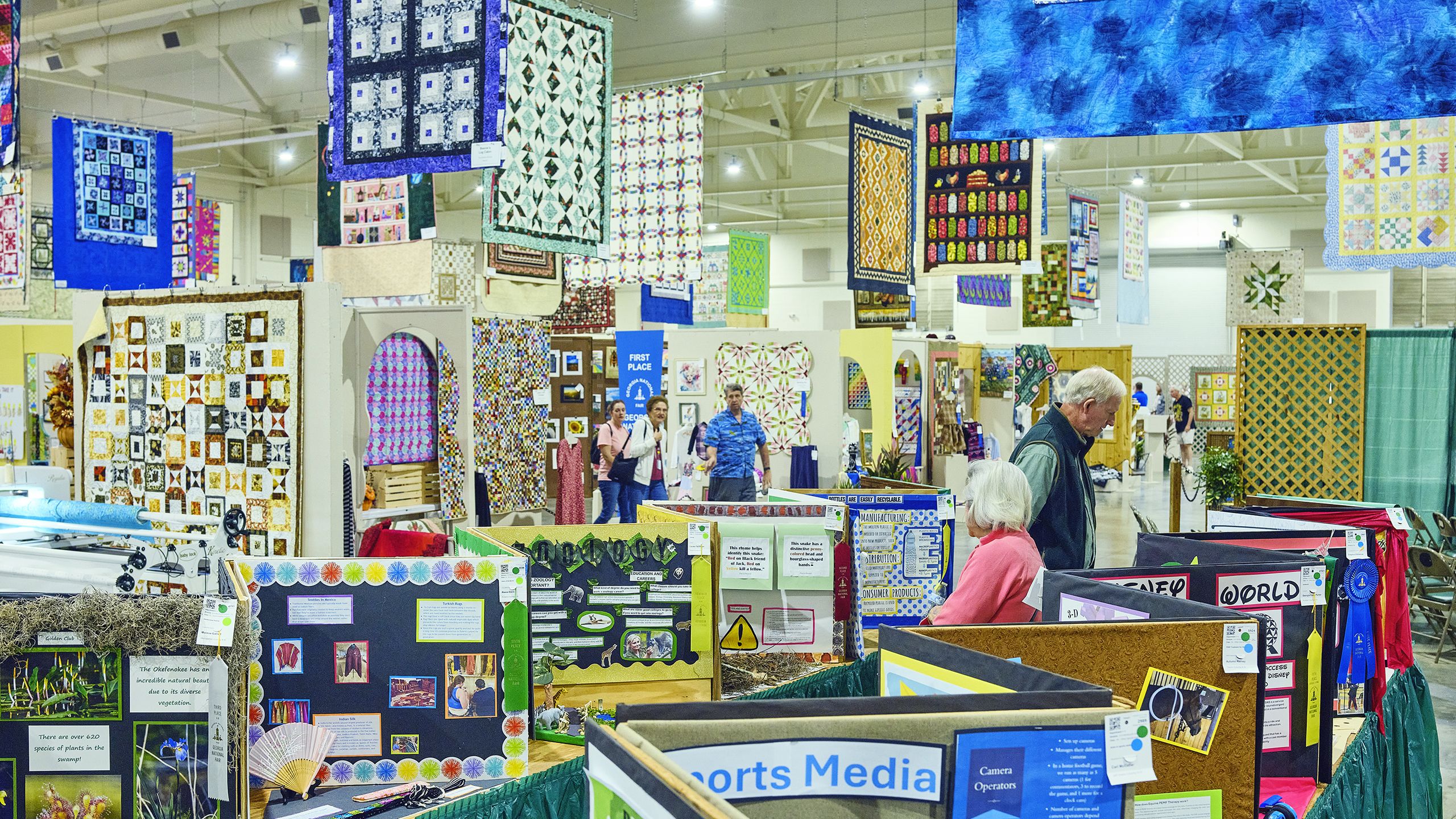 Fair goers visit the exhibit hall at the Georgia National Fair crowded with colorful booths and handmade quilts hanging from the ceiling and walls.