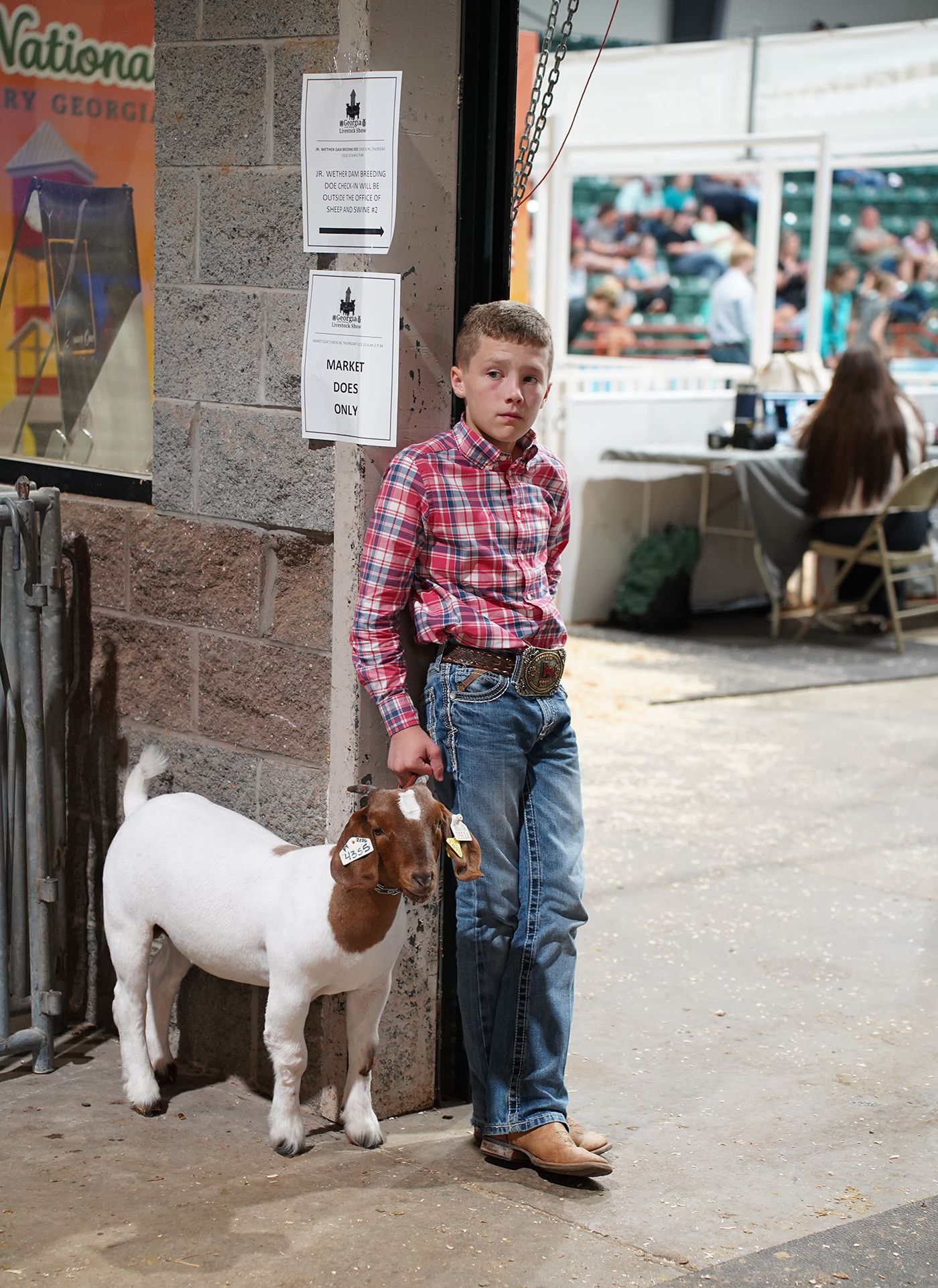 A 4-H student in jeans and a plaid shirt leans against a doorway holding the reins of a white and brown goat,  waiting for his turn to show his goat at the Georgia National Fair. 