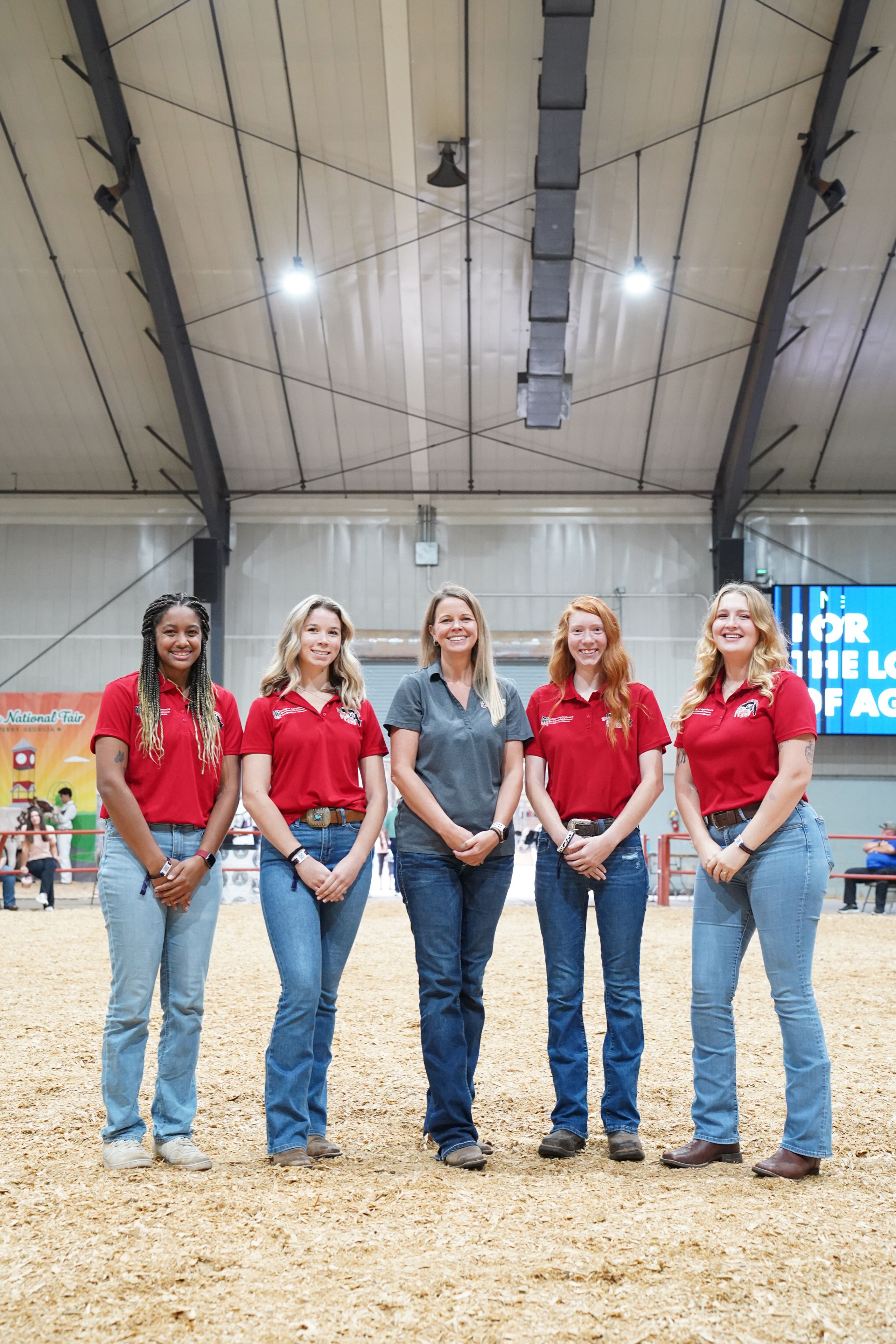 Group of five women standing in a livestock show arena at a fairground, posing for a photo. Four women wear matching red polo shirts with embroidered logos and blue jeans, while the woman in the center wears a gray polo shirt and jeans.