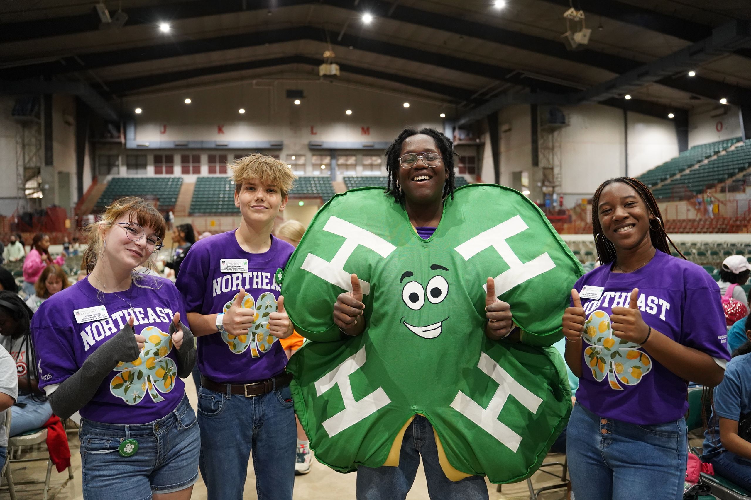 Four people in purple "Northeast" shirts pose with thumbs up; one wears a large green four-leaf clover costume.