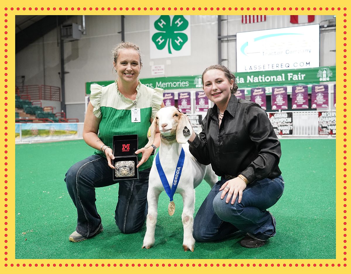 A woman in a green anad white shirt kneels displaying the belt buckle award for Goat Master Showmanship to a yound womand in a black button up shirt and jeans with her hand on the winning goat, which wears a medal around its neck. 