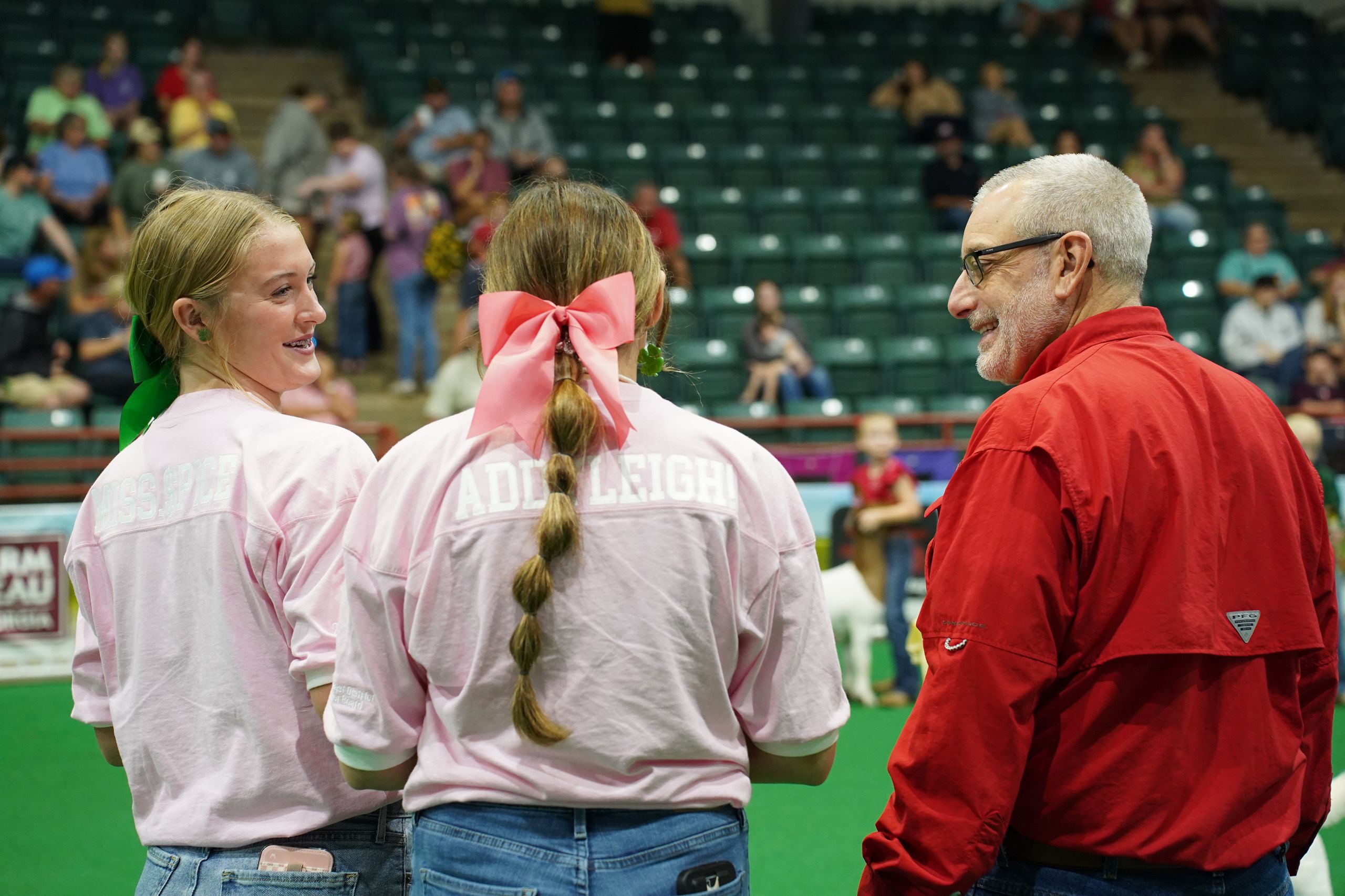 Two young women in pink t-shirts and jeans talk to a man in a red shirt in a show ring at the Georgia National Fair. 