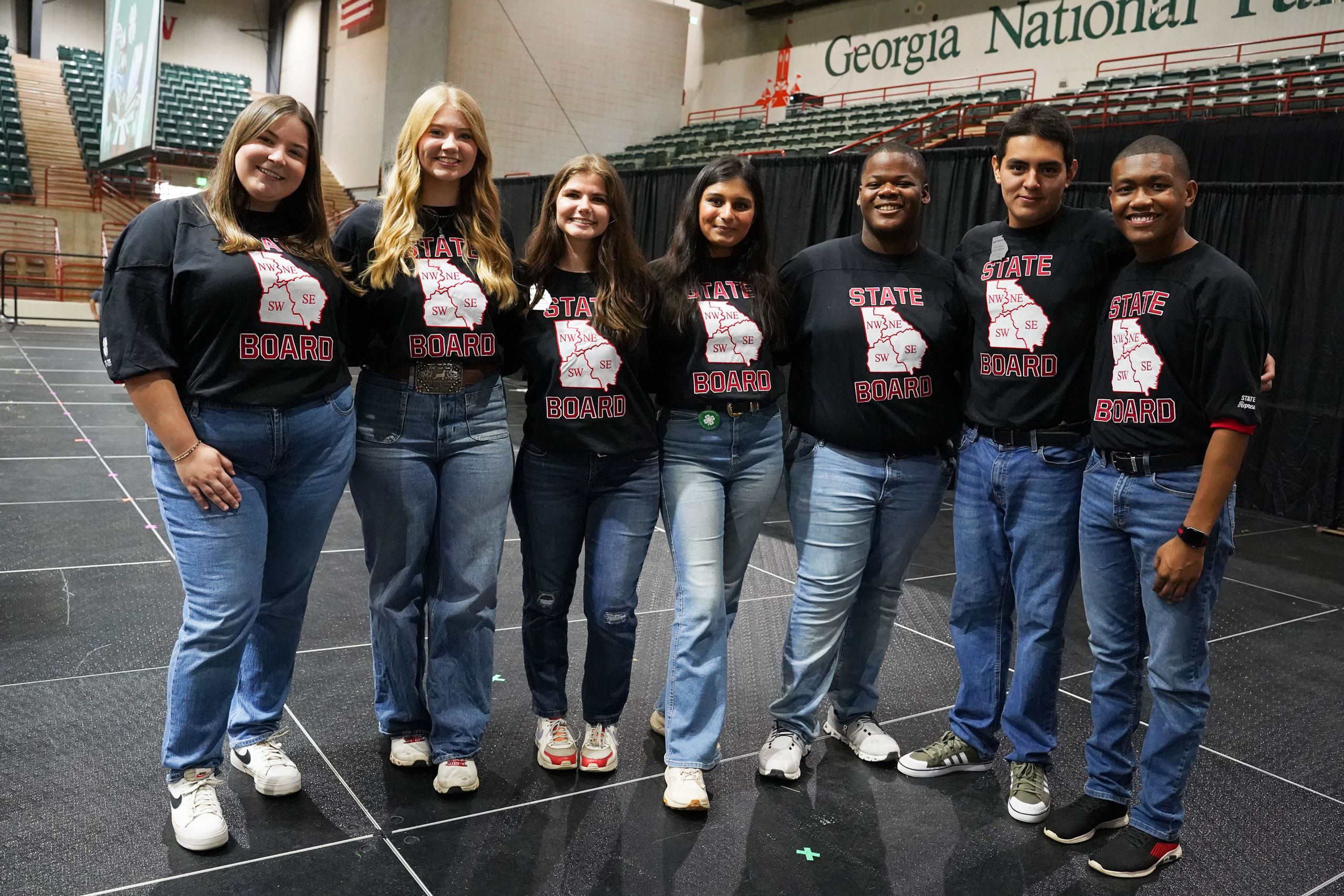 Seven people in matching black "State Board" shirts with Georgia map design stand together in an arena.