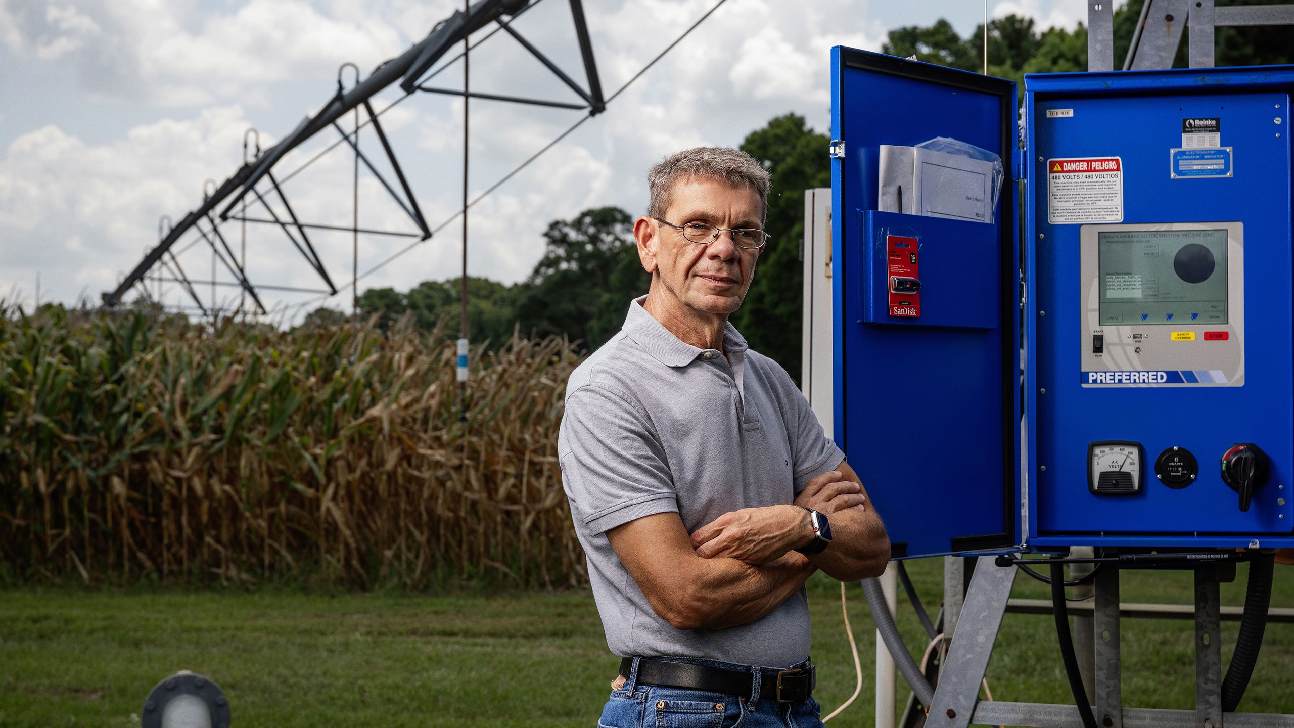Middle-aged man wearing glasses, a gray polo shirt, and a smartwatch standing with arms crossed in front of a large blue irrigation control panel outdoors. Behind him is a cornfield with tall, dry stalks and a large pivot irrigation system extending across the field.