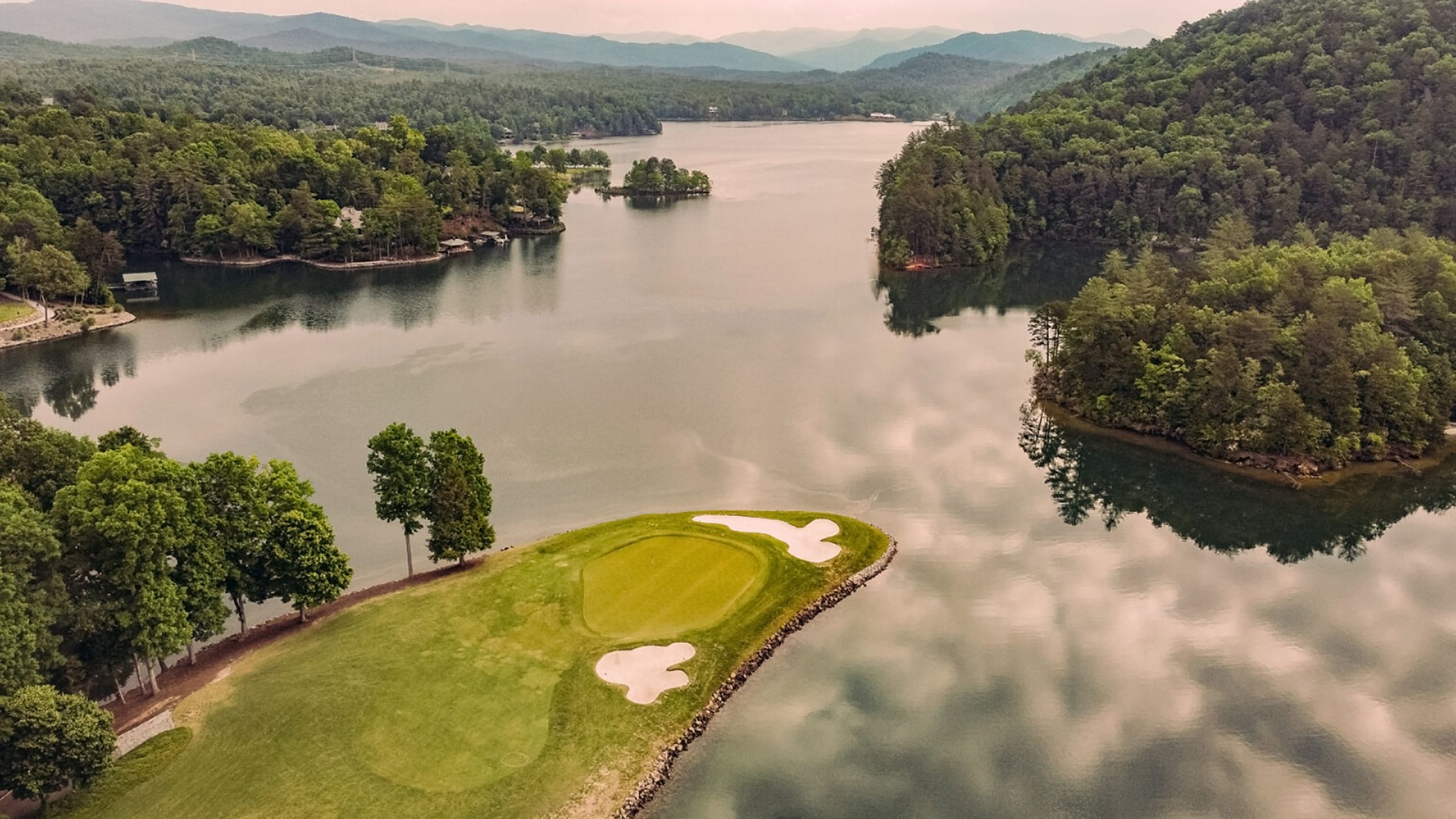 An aerial view of a golf course surrounded by water, with lush green fairways and sand bunkers, set against a backdrop of dense forests and distant mountains