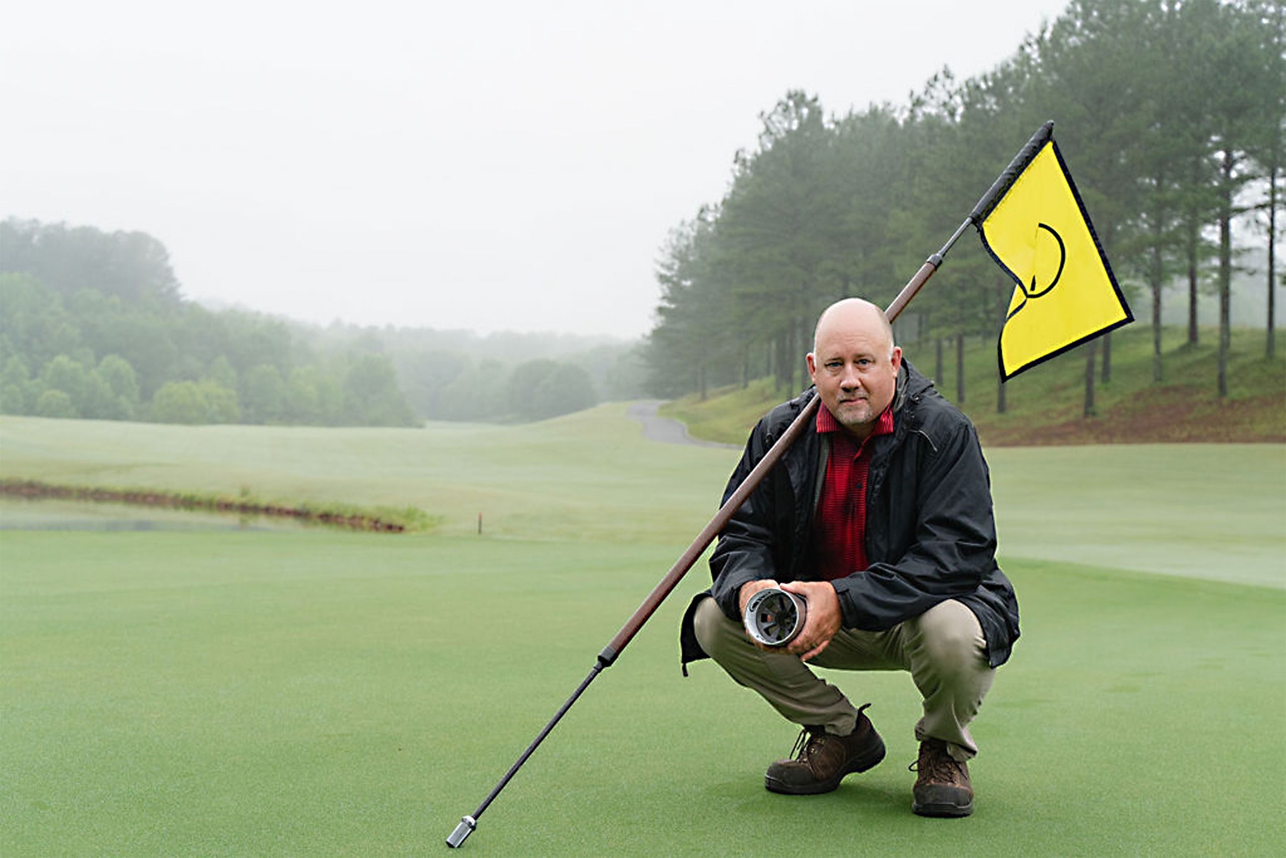 Jim Evans kneels on a golf course, holding a hole cup, with a yellow flag in the background, set against a misty, serene landscape.