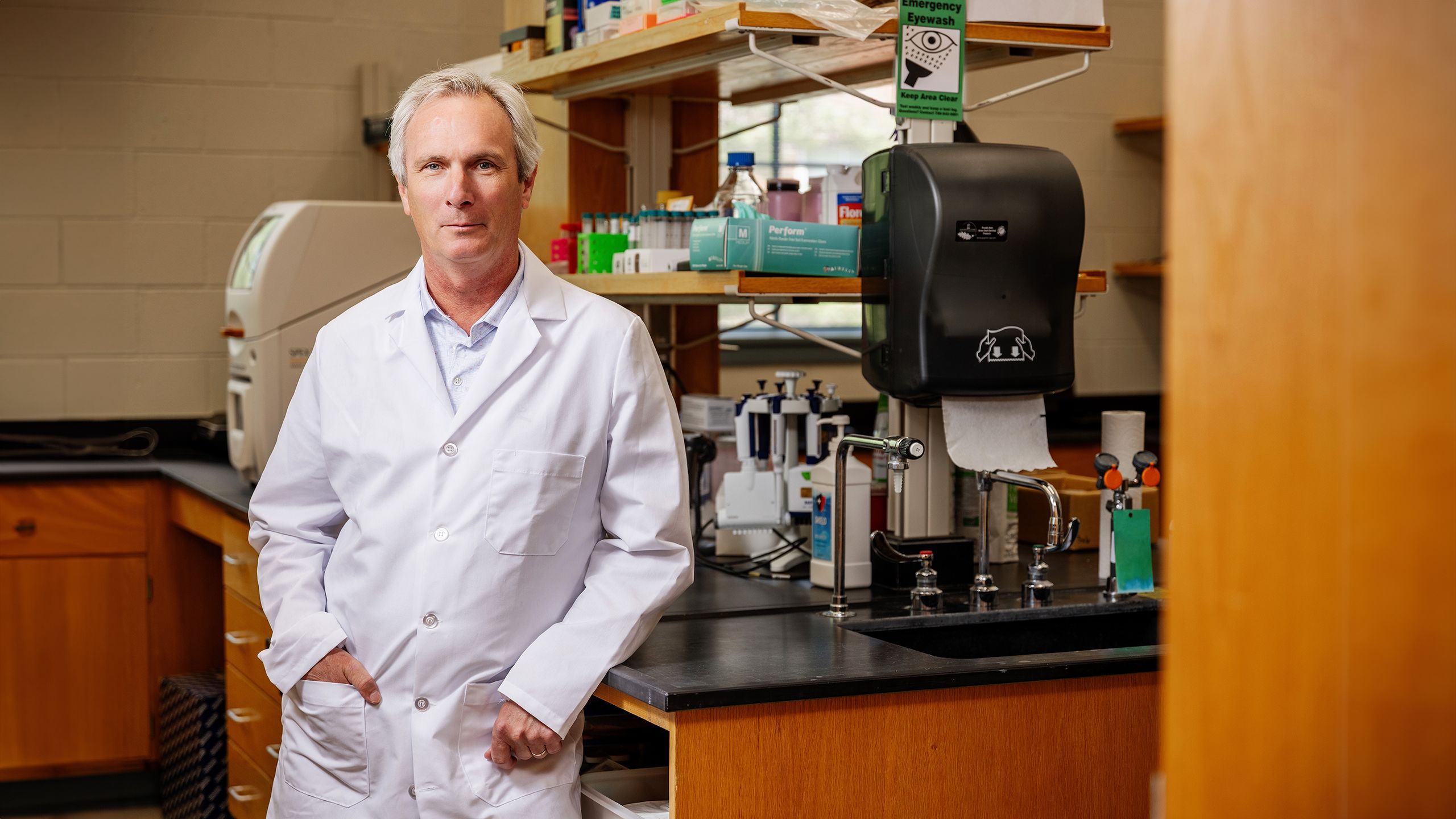 Man in a white lab coat stands in a laboratory with scientific equipment, shelves, and a sink in the background.