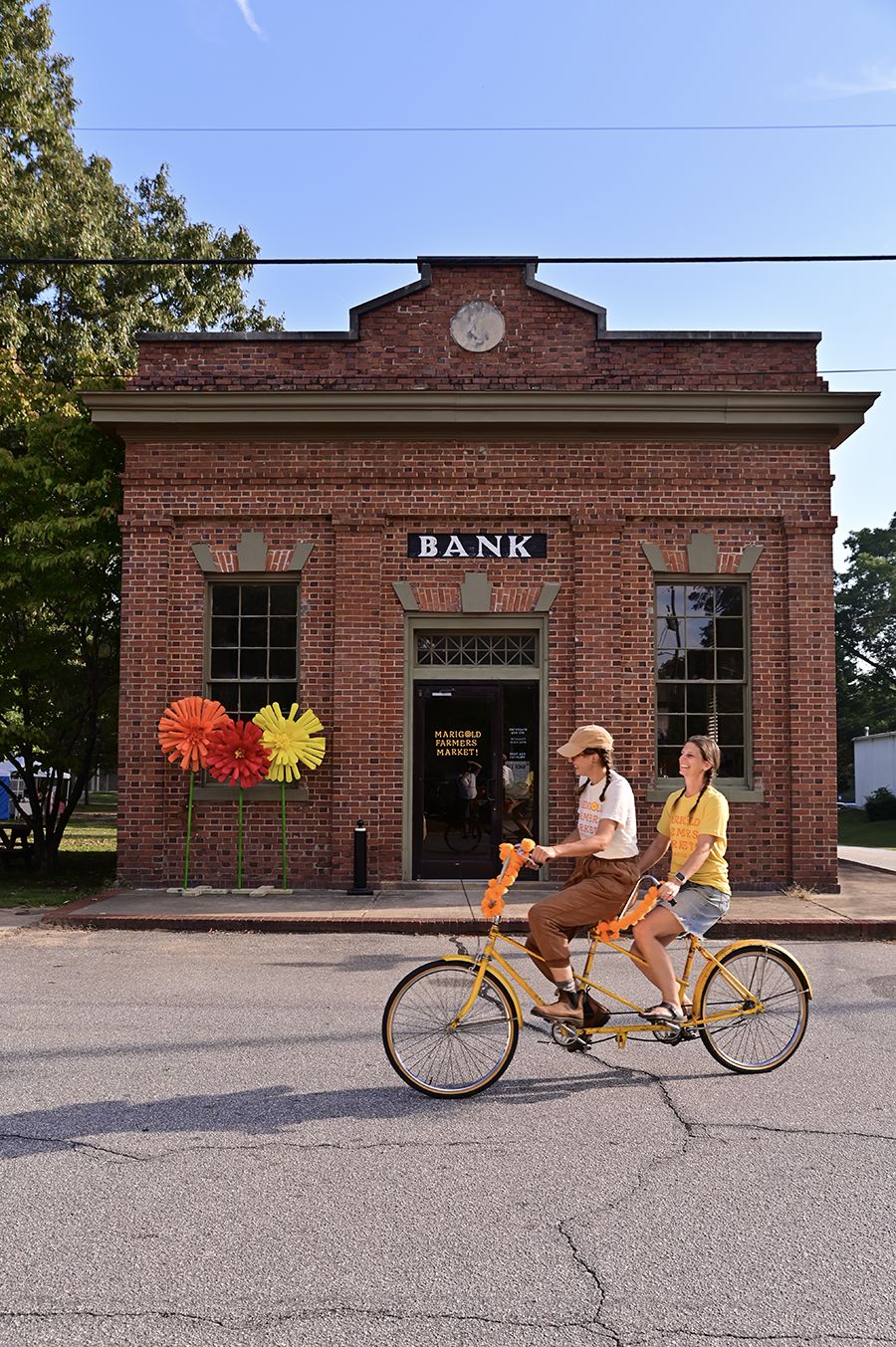 Two women ride a yellow tandem bicycle decorated with orange flowers past a historic brick building marked “BANK,” with large colorful flower decorations and a door labeled “Marigold Farmers Market.”