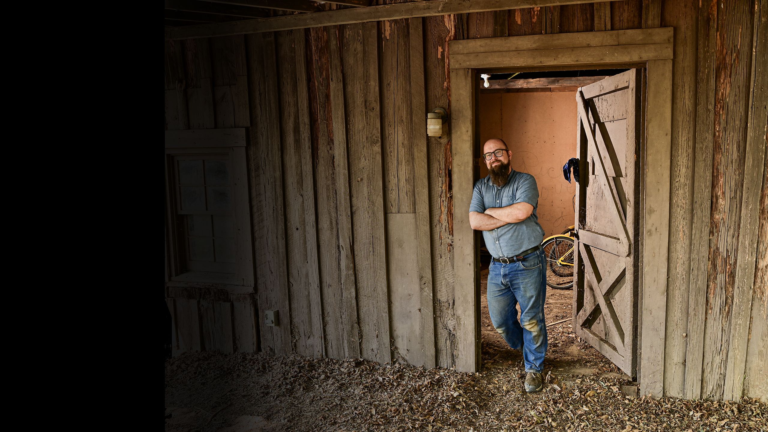 A bearded man in a denim shirt and jeans stands with arms crossed in the doorway of a rustic wooden building, smiling confidently.
