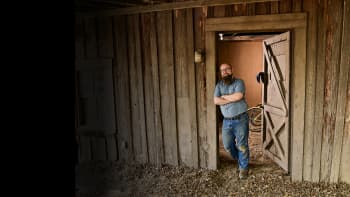 A bearded man in a denim shirt and jeans stands with arms crossed in the doorway of a rustic wooden building, smiling confidently.