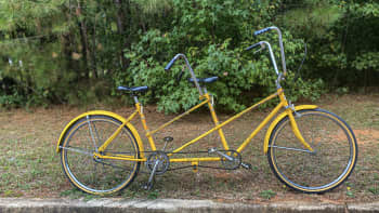 A bright yellow restored tandem bicycle with high handlebars is parked on a path in front of green foliage.