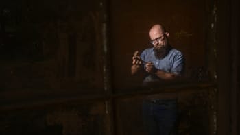 A bearded man wearing glasses and a denim shirt carefully inspects or shapes a curved metal tool under focused lighting.