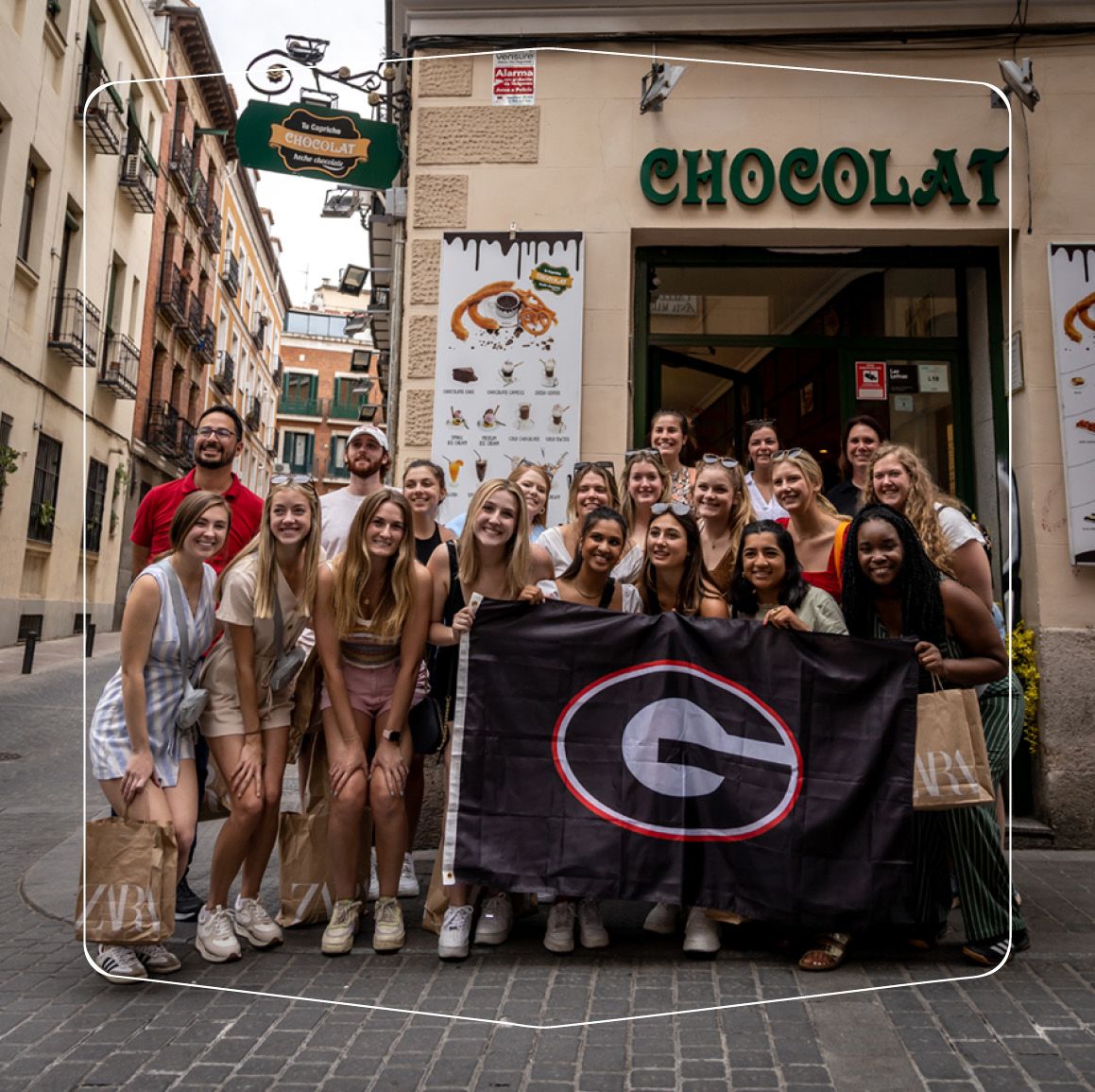 A group of UGA students hold up a UGA flag in front of a chocolate shop in Spain.
