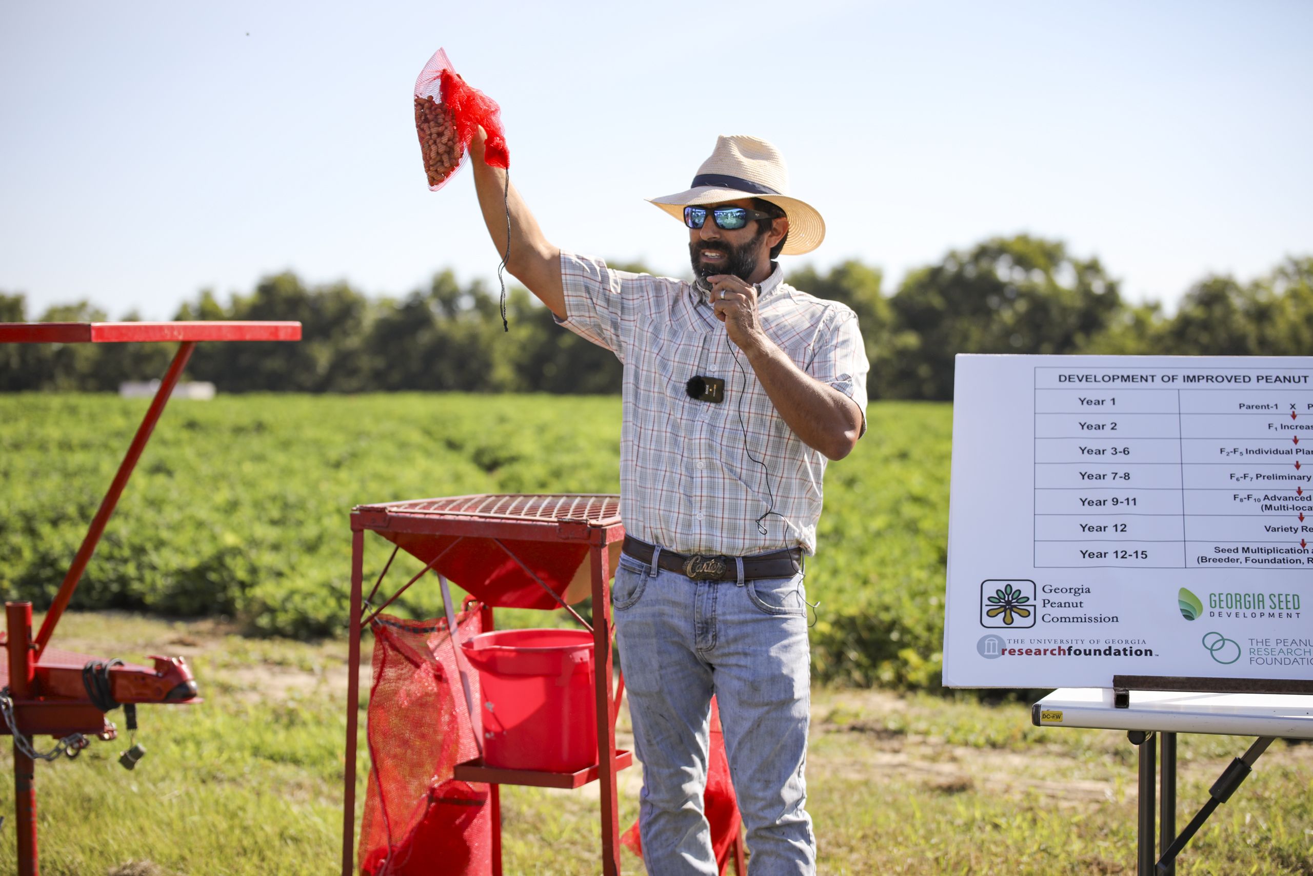 Nino Brown, an assistant research scientist stands in front of a peanut field surrounded by research equipment and holding a red net bag of peanuts in his raised right hand.