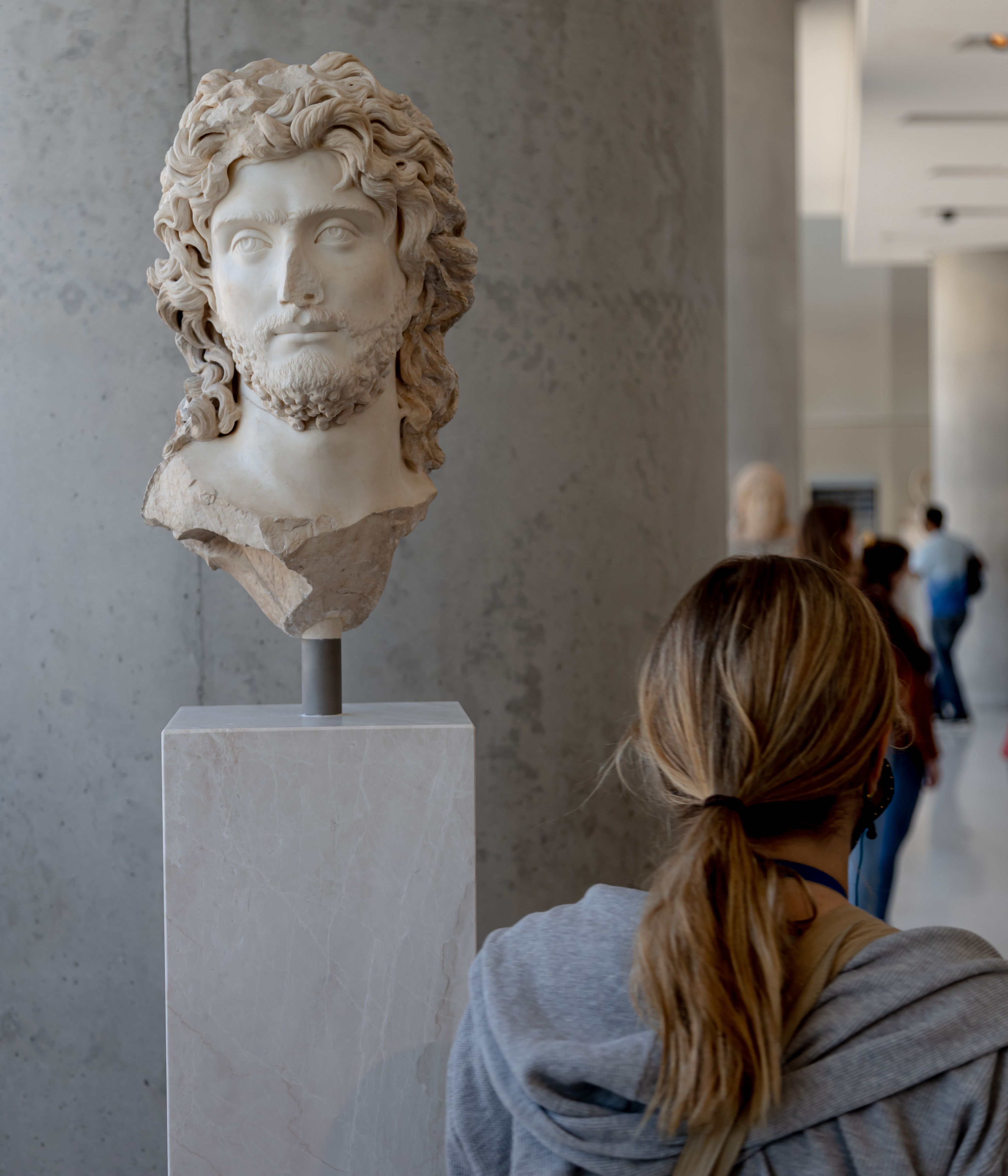 A photo from behind of a blonde, ponytailed young woman as she faces a marble bust on display in the Acropolis.