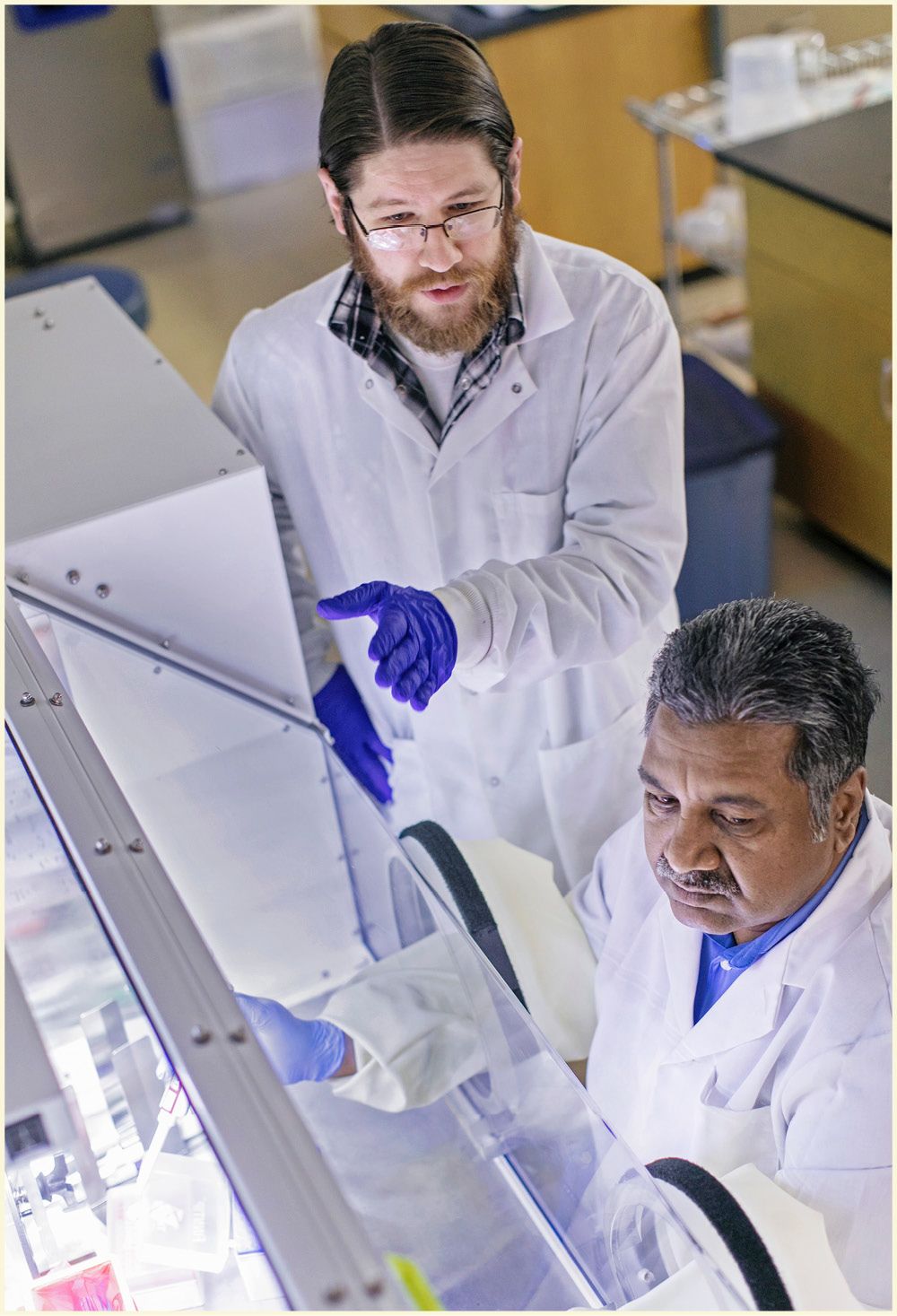 Two people in lab coats stand in a lab looking into a fume hood