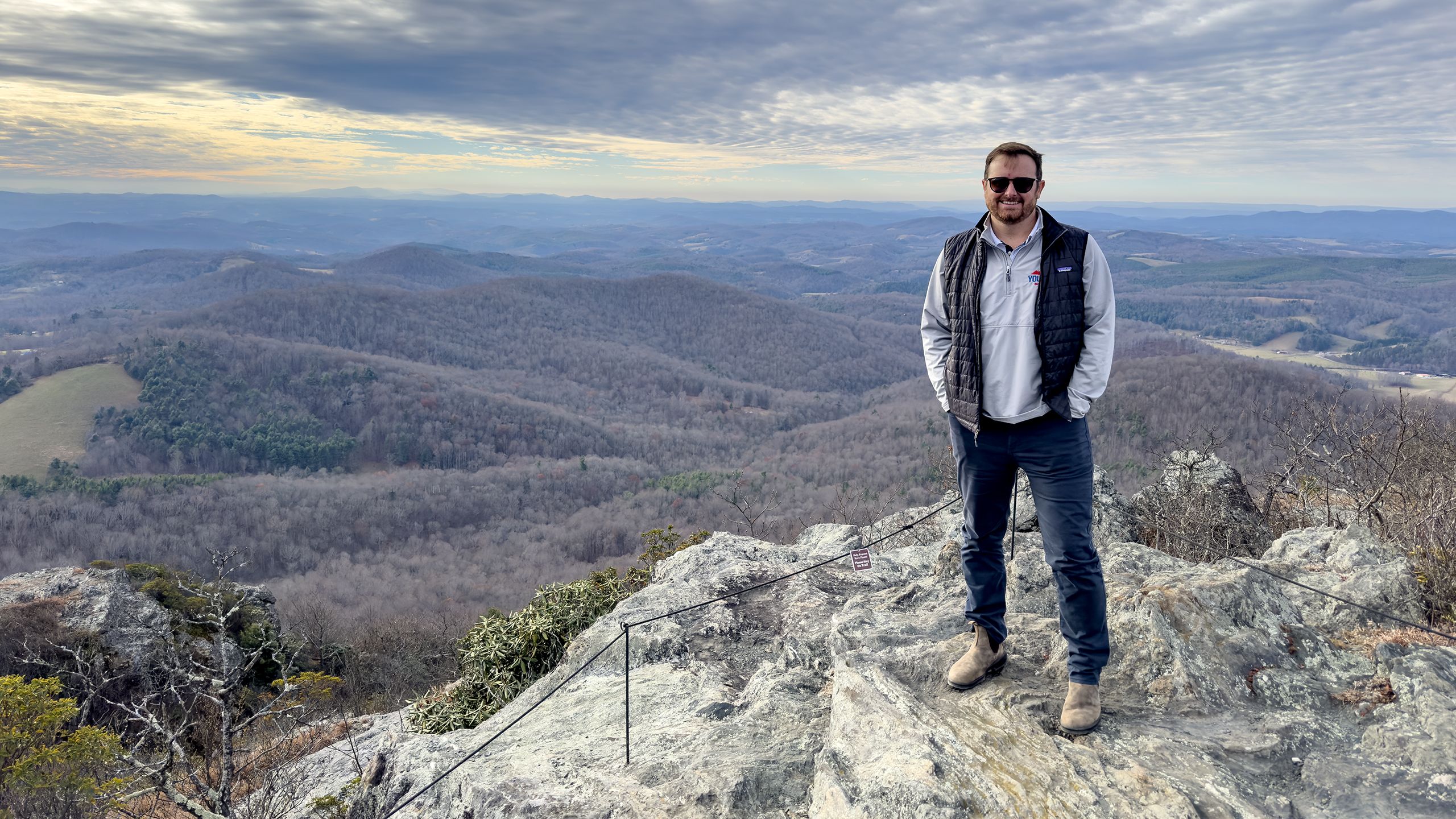 A man stands on top of a mountain in casual attire and sunglasses.