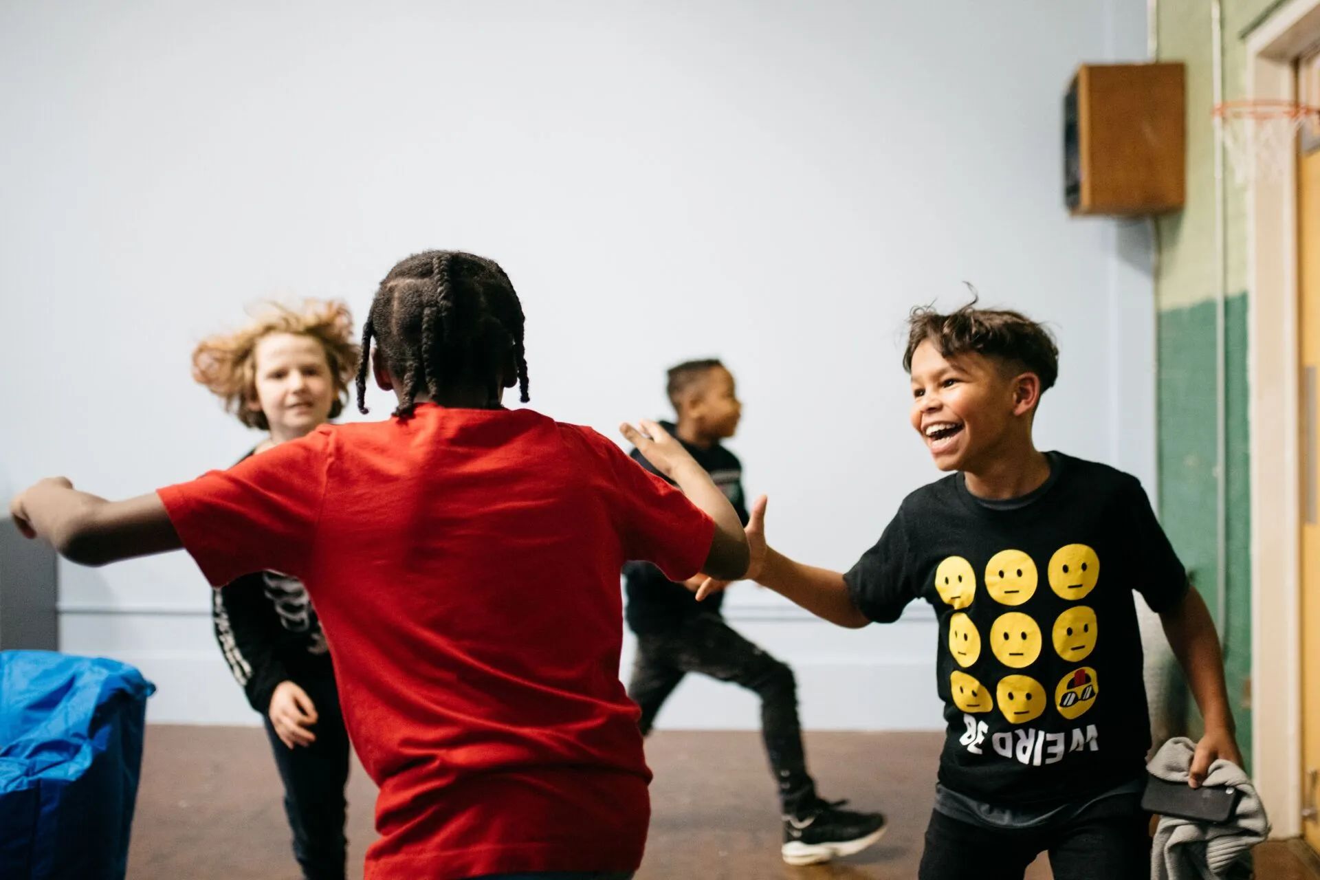 Several children playing and running around. In the foreground, a young boy wearing a black shirt with smiley faces on it smiles at another young boy in a red shirt.