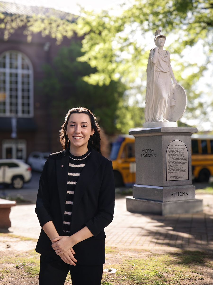 A woman in a black blazer and striped top stands smiling in front of a statue of Athena with a school building and trees in the background.
