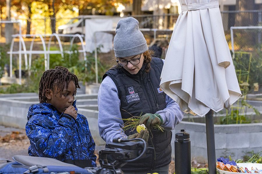 A woman in a UGA Extension vest and gray beanie teaches a young student about plants outdoors, holding a small plant while the young student attentively.