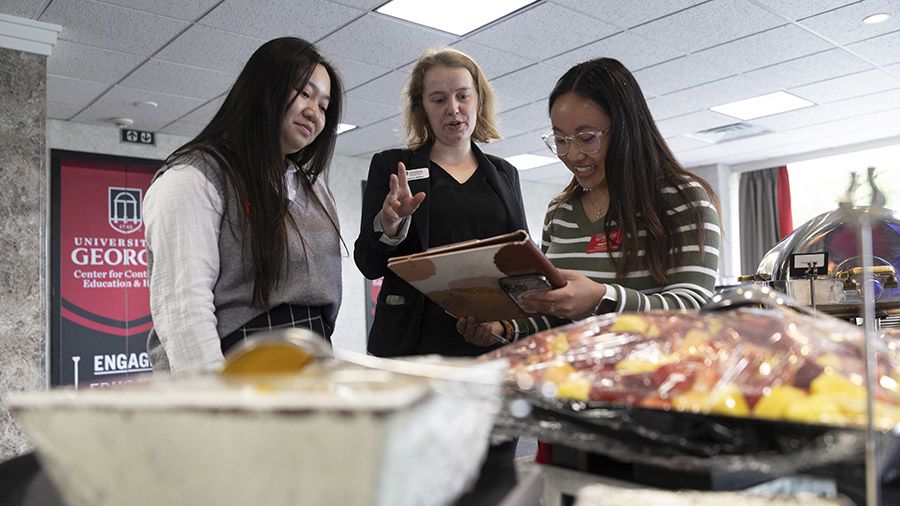 Three women review a buffet setup at the University of Georgia Center for Continuing Education and Hotel, with one holding a tablet and gesturing as the others listen.