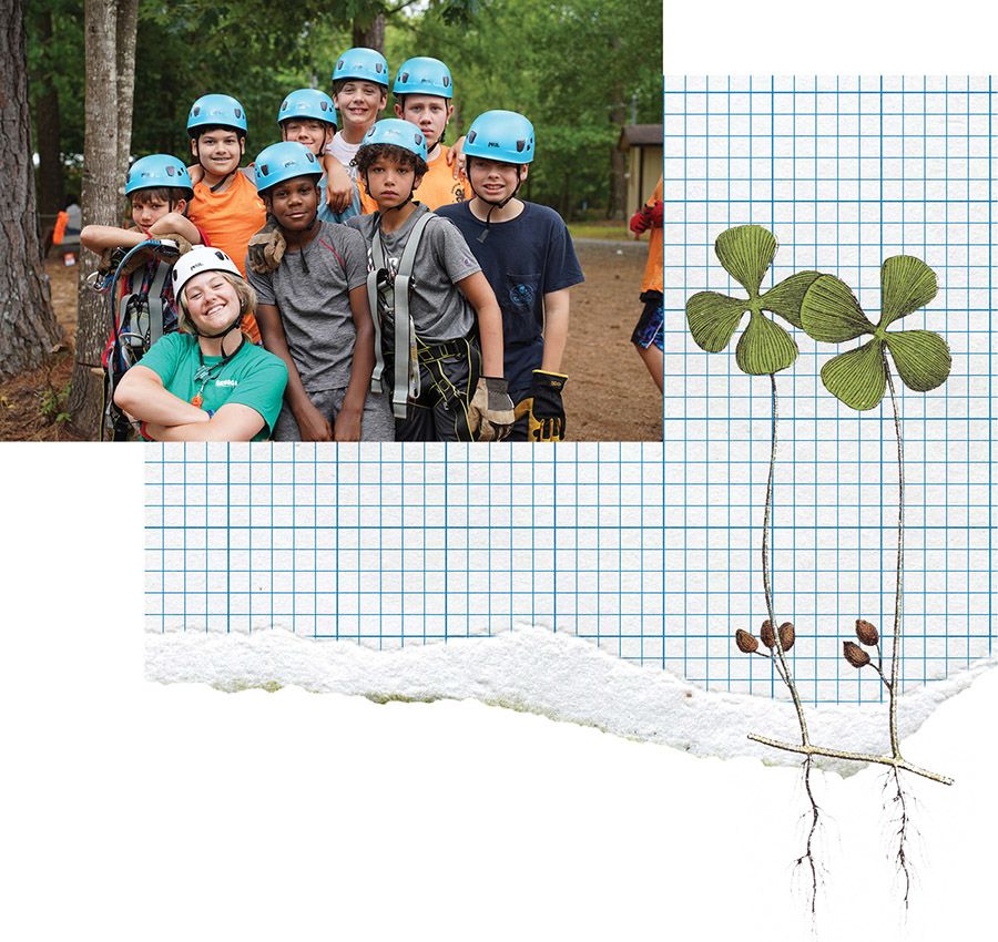 A group of children wearing blue helmets and harnesses pose in a wooded outdoor setting, with a smiling adult in a green shirt kneeling in front.