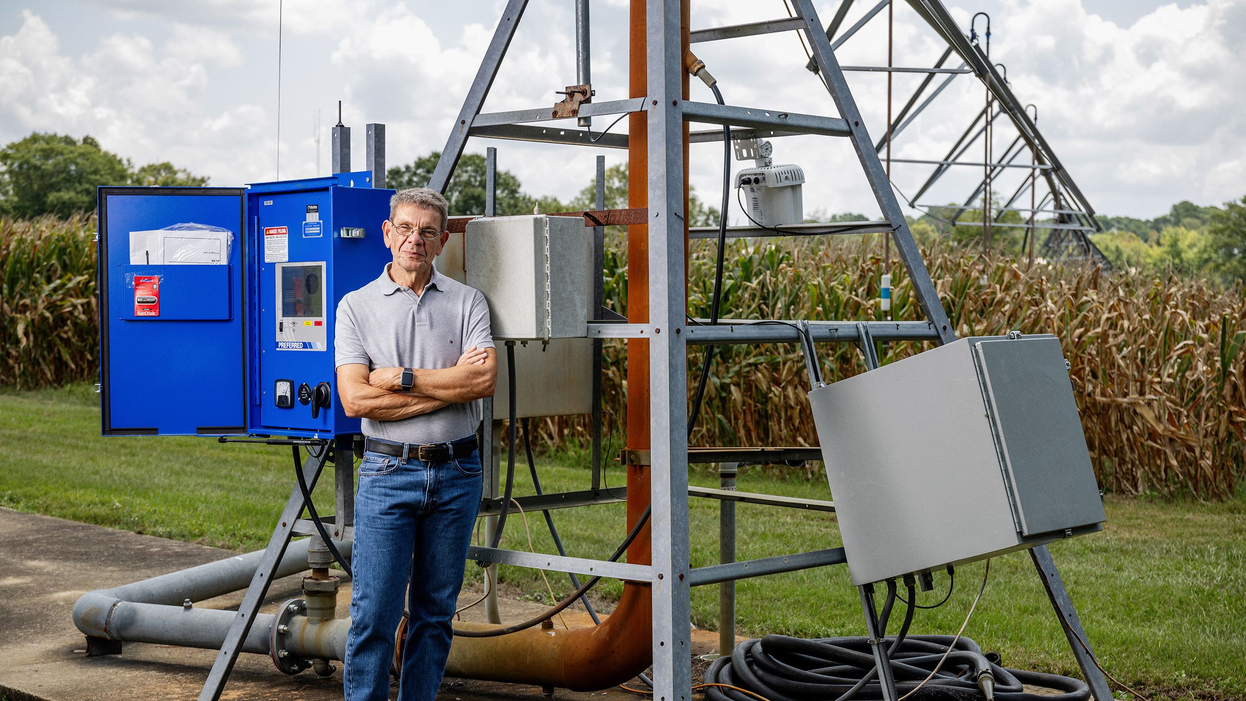 George Vellidis stands with his arms crossed in front of a blue control box and irrigation equipment, set against a backdrop of a cornfield under a partly cloudy sky.